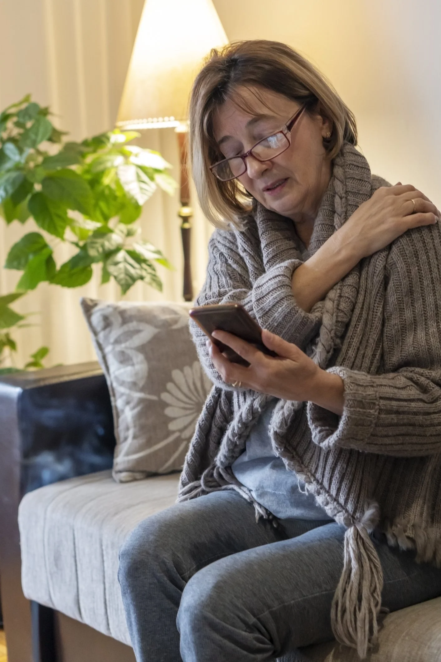 A woman with glasses sitting on a beige couch, looking at her phone with a concerned expression, hand on shoulder, in a cozy room with a lamp and plant.