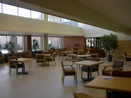 An empty, modern lobby or lounge area with several tables and chairs, large windows, potted plants, and a reception counter in the background.