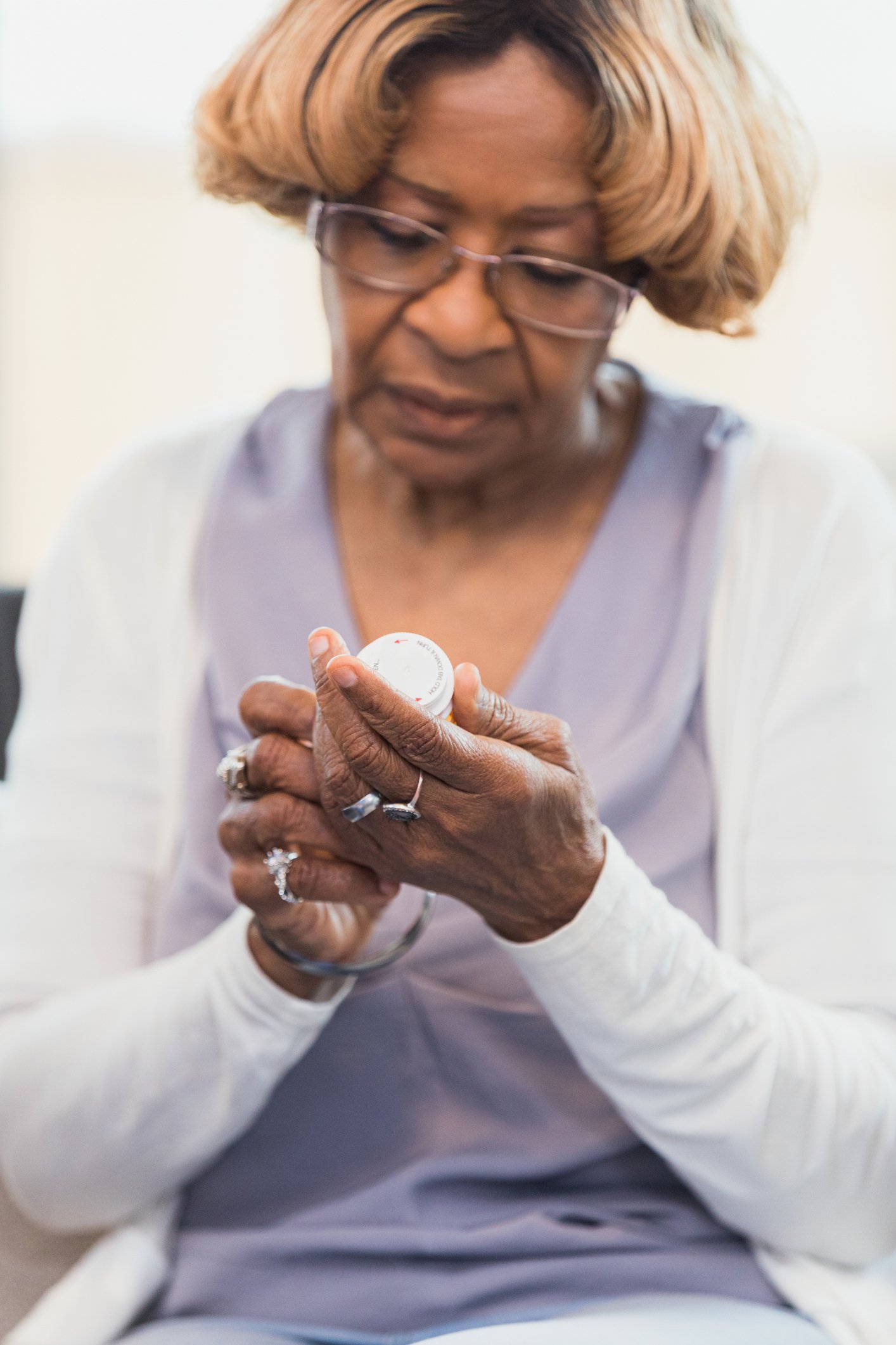 An elderly woman with glasses, wearing a lavender top and white cardigan, looks at a pill bottle she is holding in her hands.