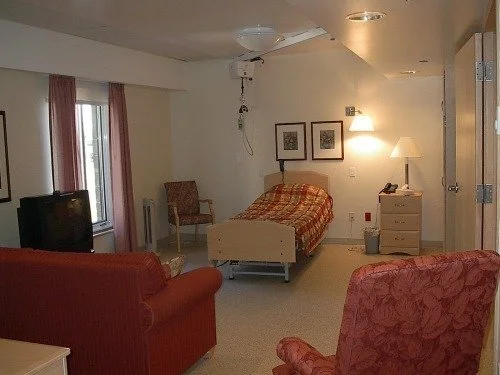 A cozy hotel room with a single bed, a red upholstered armchair, a TV, and a window with pink curtains. The room has a nightstand, lamps, and framed artwork on the wall.