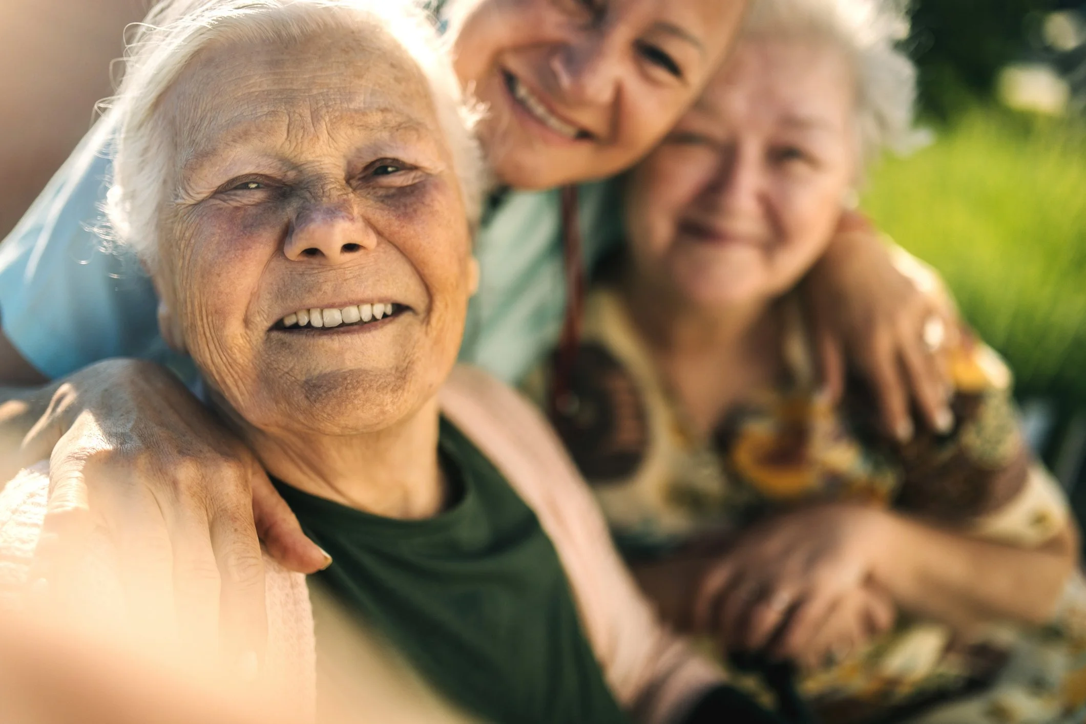 Three elderly people smiling and sitting close together outdoors, with one person in the foreground and two in the background