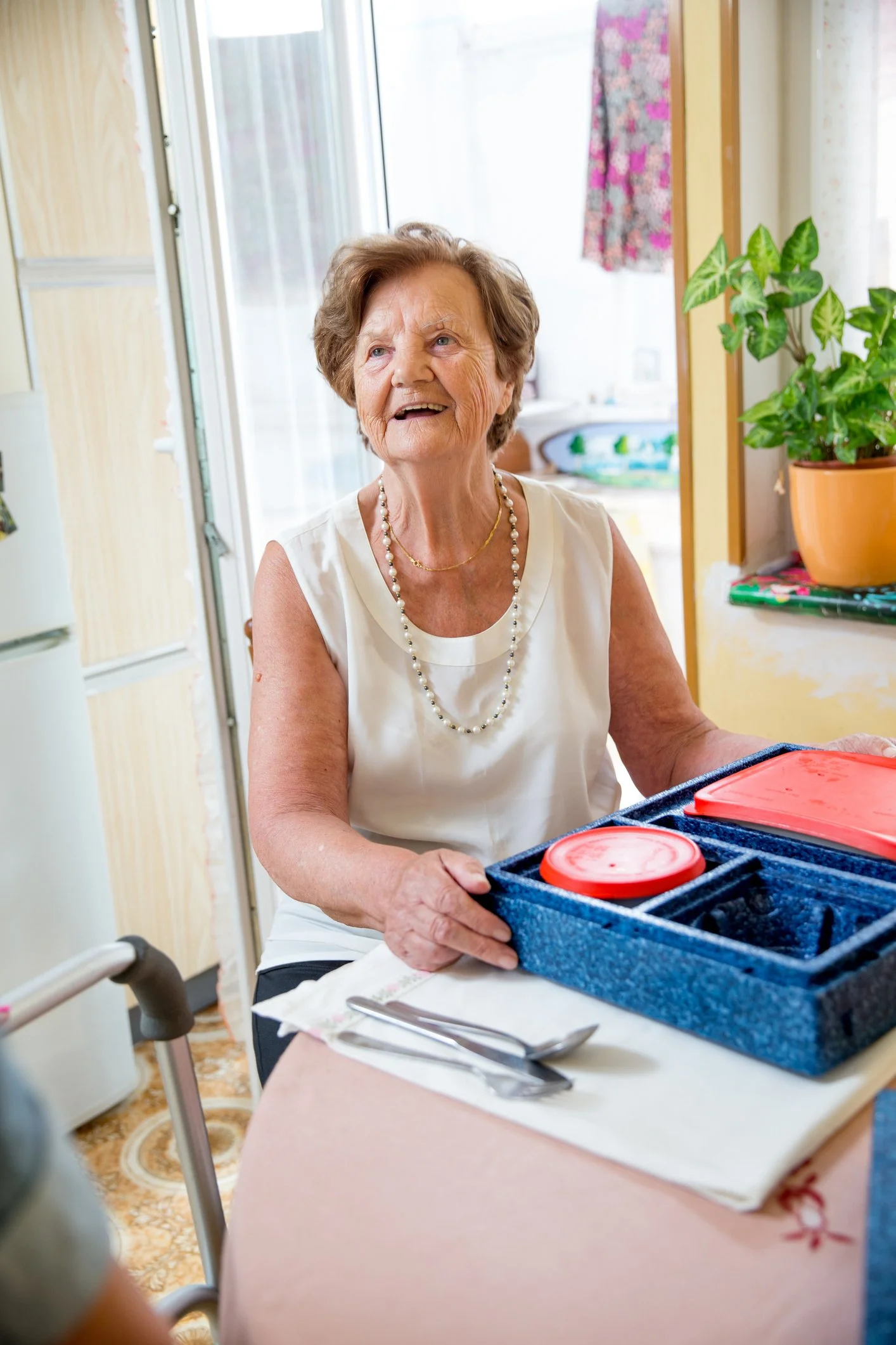 An elderly woman sitting at a table in a bright room, holding a blue container with red lids, smiling and looking up.