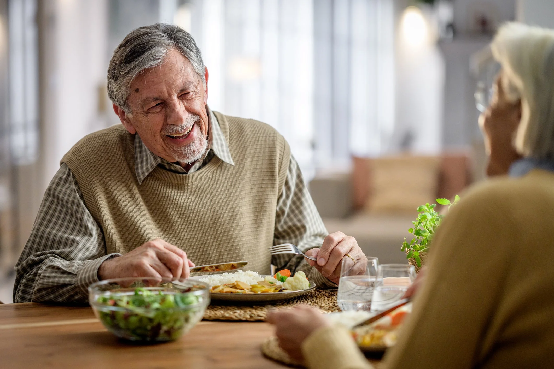 Two elderly people, a man and a woman, enjoying a meal and smiling at each other at a dining table in a bright, cozy room.