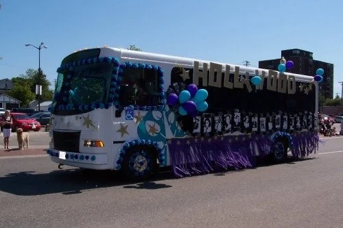 Float decorated for a parade, featuring the word 'Hollywood' and images of celebrities, with balloons and purple fringe, driving down a street.