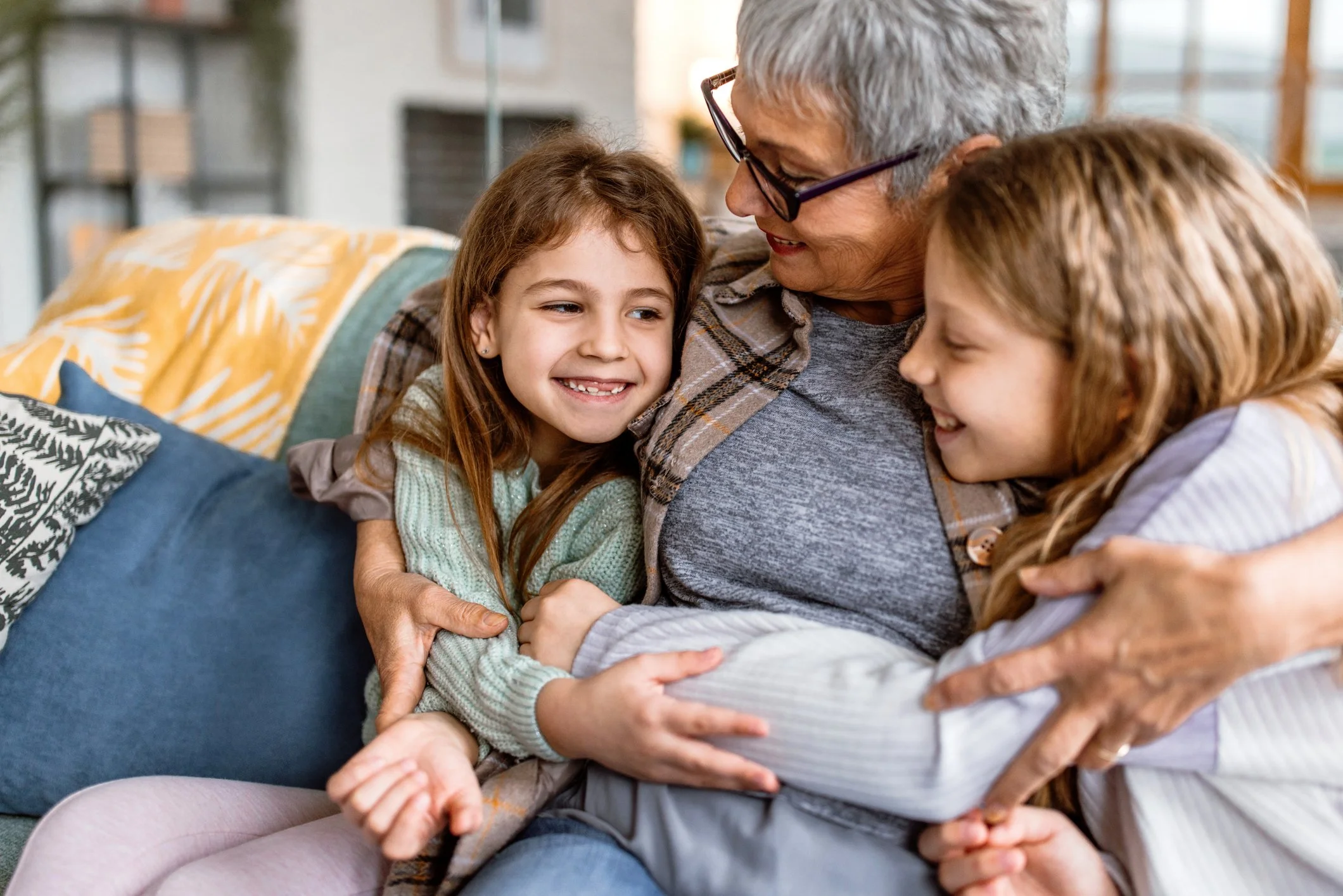 Elderly woman with gray hair and glasses hugging two young girls with brown hair, all smiling and embracing each other on a couch in a cozy living room.