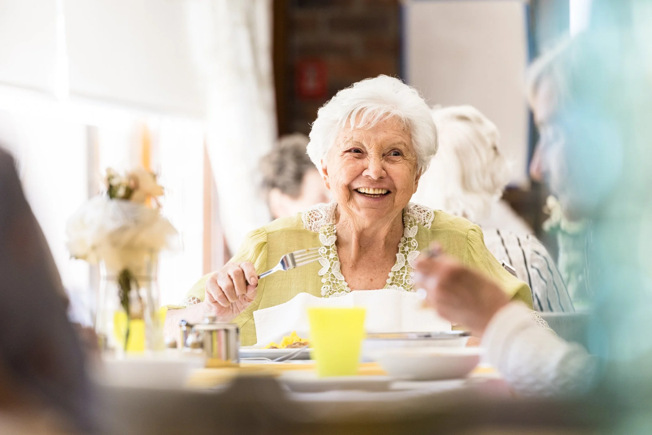 An elderly woman with white hair smiling and holding a fork while sitting at a table during a meal with other people.