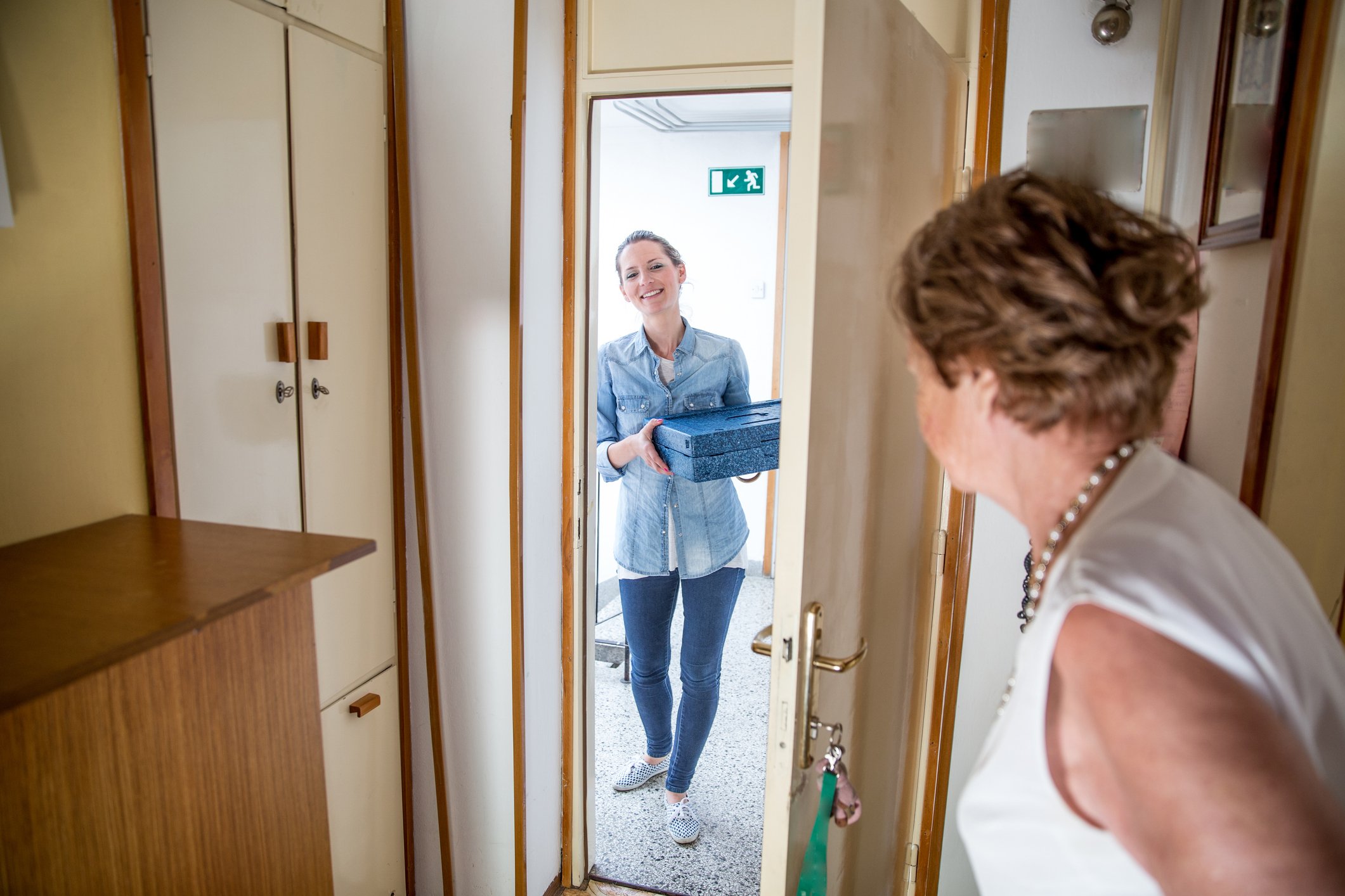 A woman standing at a doorway receiving a gift box from another woman inside a home.