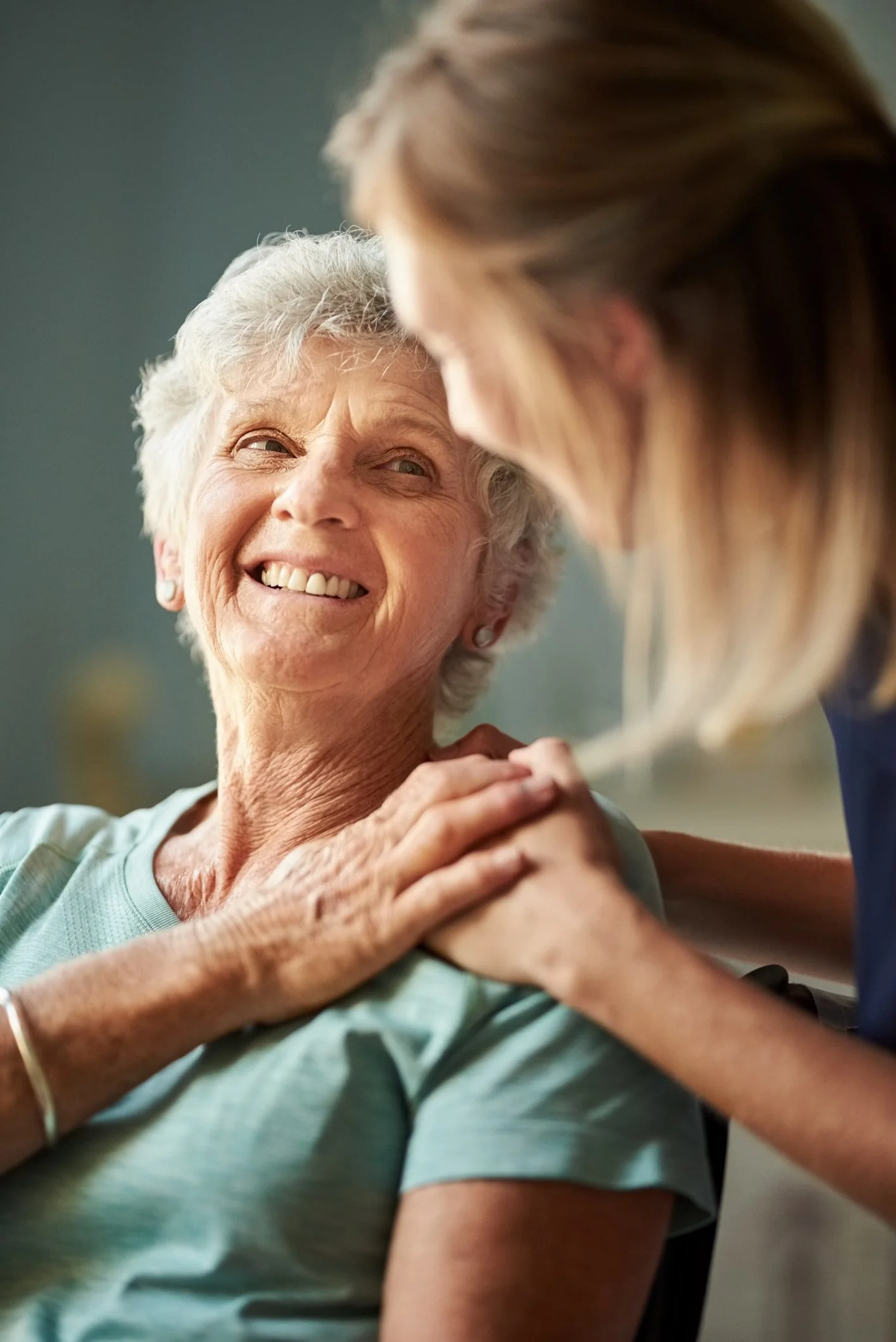 A healthcare worker smiling and holding the shoulder of an elderly woman, who is smiling back warmly.