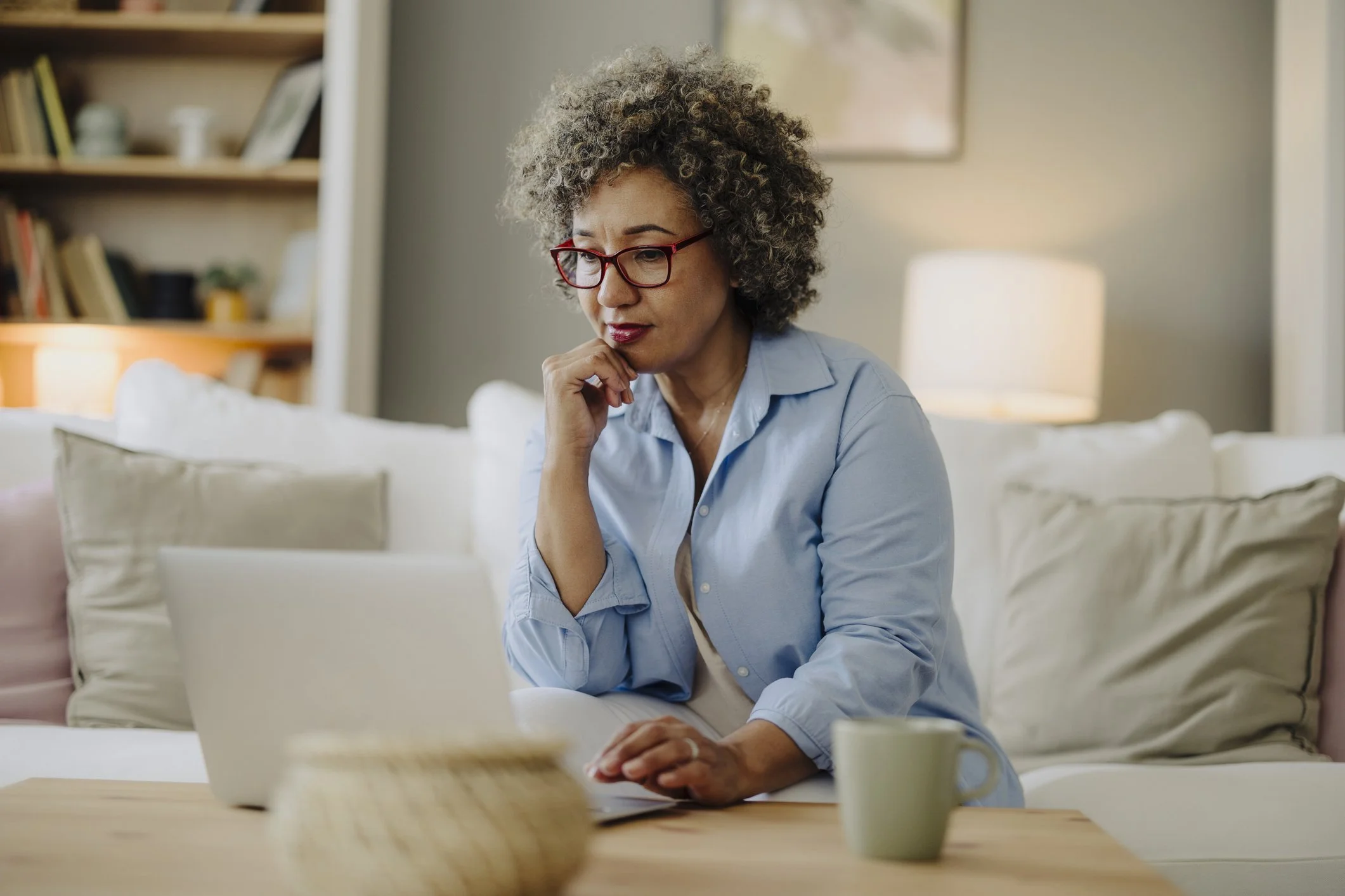 A woman with curly gray hair and red glasses working on a laptop at home, sitting on a sofa with pillows, with a bookshelf and a lamp in the background.