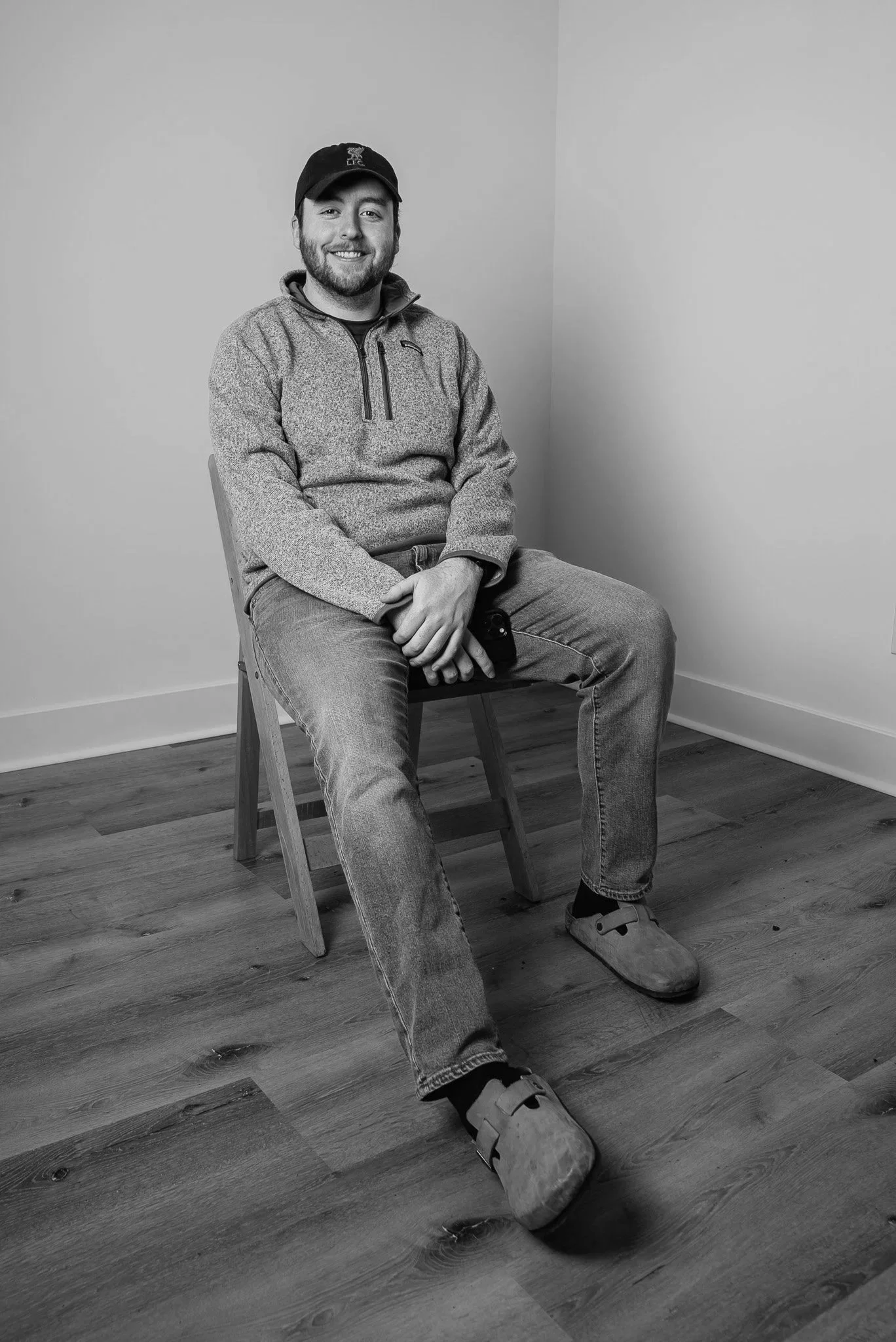 A smiling man sitting on a wooden chair in a corner of a room, wearing a cap, a fleece jacket, jeans, and slippers, holding a phone in his lap.