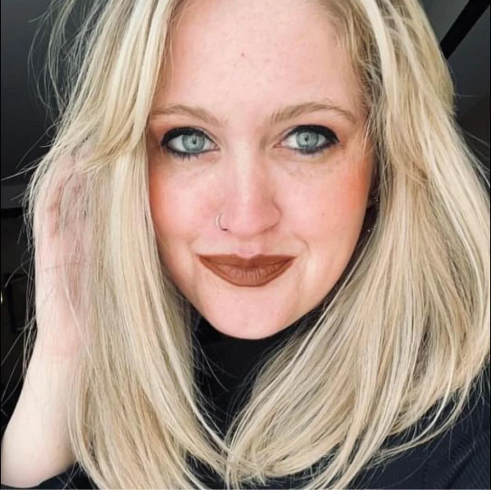 A woman with long blonde hair, blue eyes, a nose piercing, wearing dark lipstick, looking at the camera, smiling slightly.