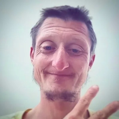 A man with short hair and a slight beard making a peace sign with his fingers, smiling softly.