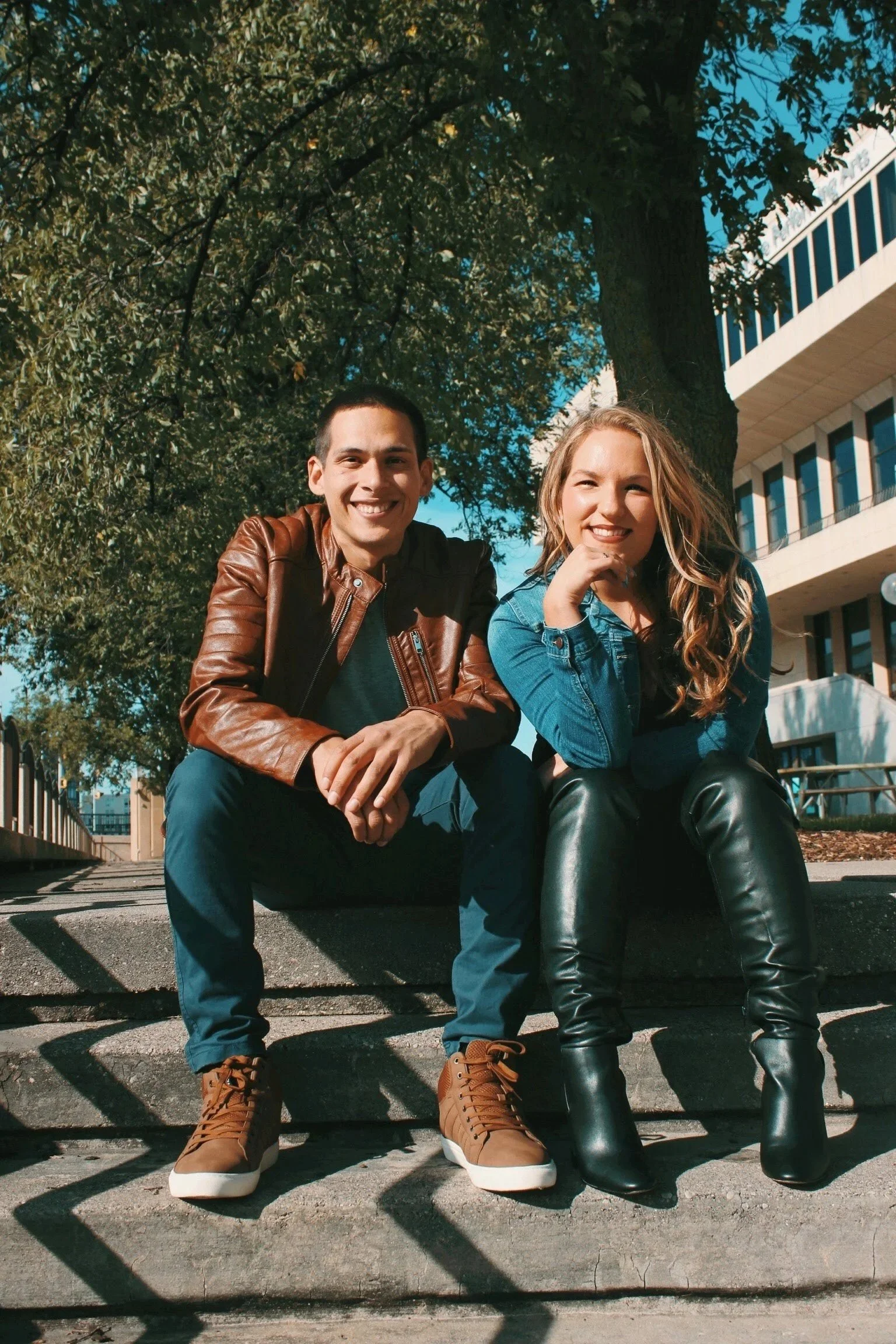 A young man and woman sitting on a concrete step outdoors under a tree, smiling at the camera. The man is wearing a brown leather jacket, dark shirt, and tan boots. The woman is in a blue denim jacket, black leather pants, and black boots. A building with multiple windows is visible behind them.