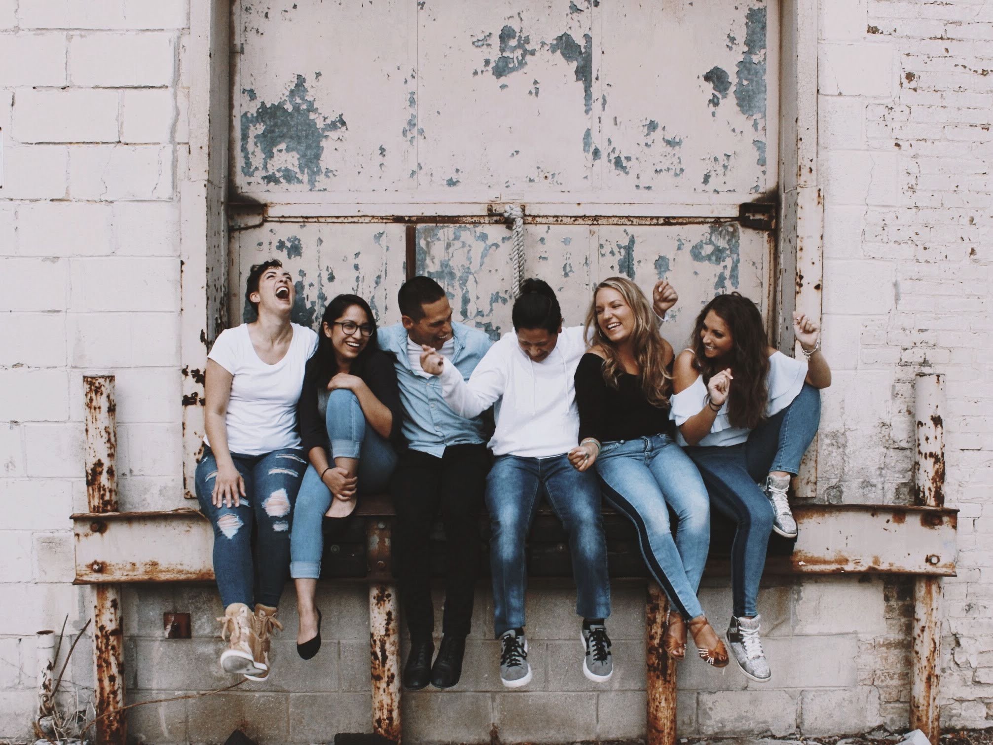 Group of seven diverse young adults sitting on an old, rusty bench against a rustic brick wall, laughing and celebrating.