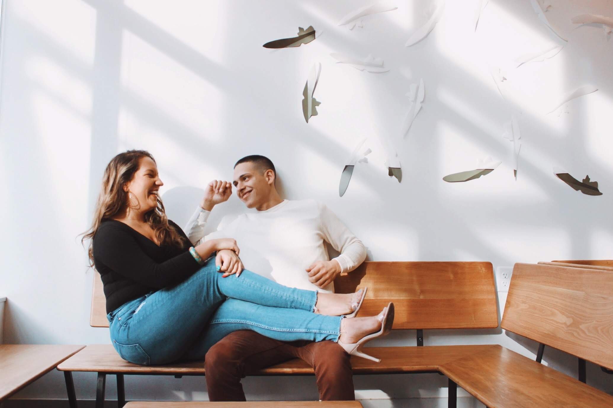 A couple sitting on a wooden bench, smiling and laughing together, with the woman sitting on the man's lap. The woman is wearing a black sweater, blue jeans, and heels, while the man is wearing a white sweater and brown pants. The background features a white wall with decorative fish-shaped mirrors and shadows cast by window blinds.