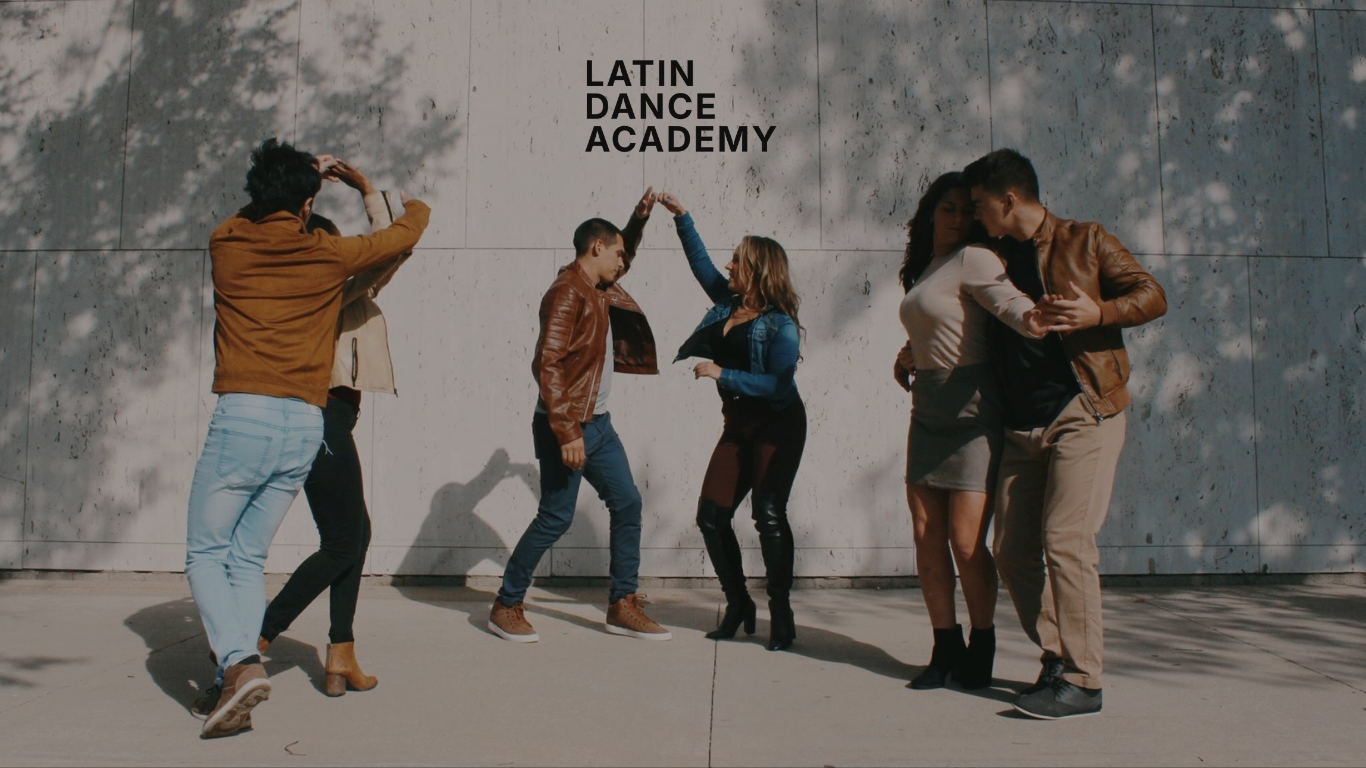 A group of six young adults dancing and socializing outdoors in front of a concrete wall with the words 'Latin Dance Academy' written on it.