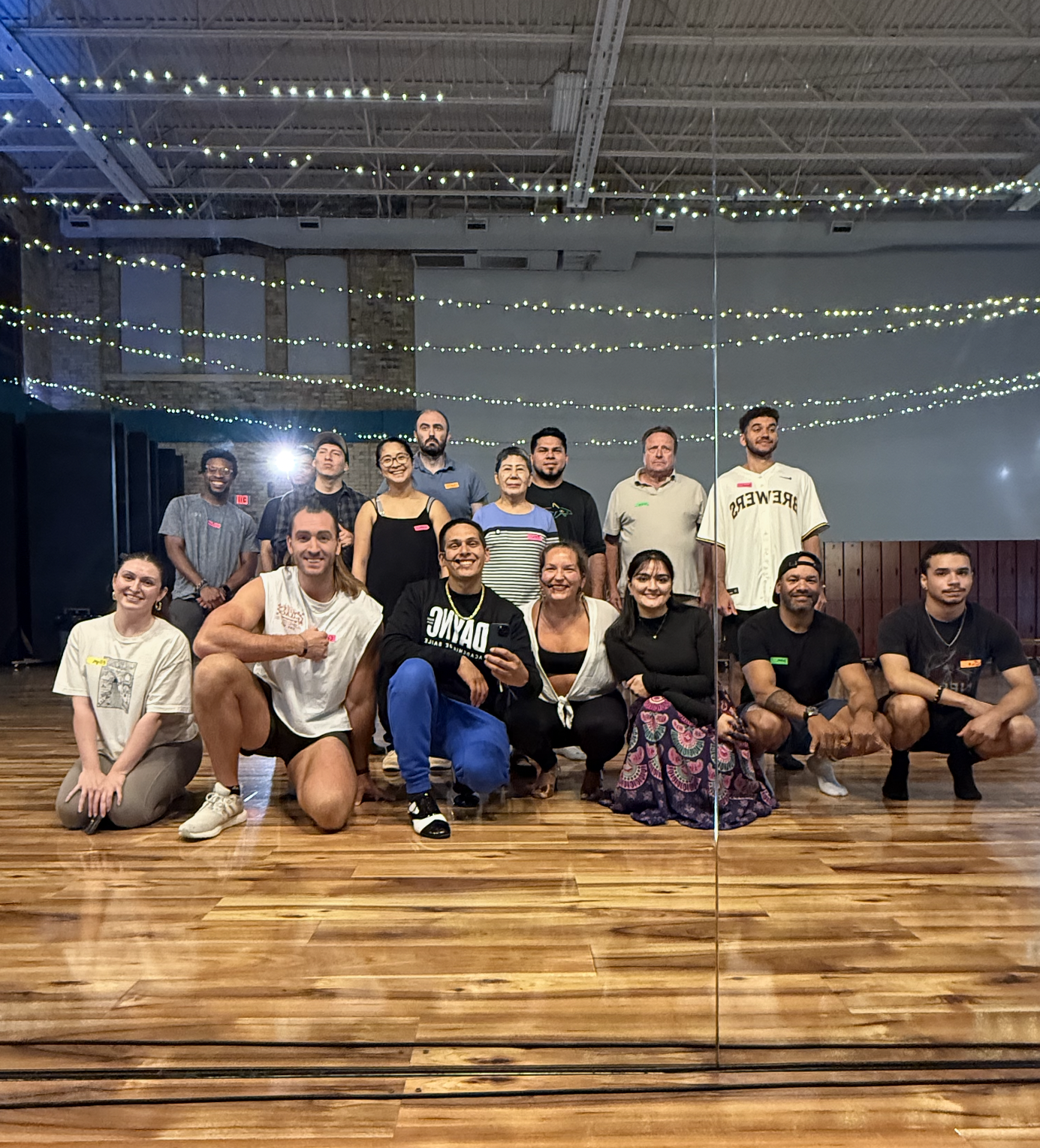 Group of people posing for a photo in a dance studio with wooden floors, string lights, and a mirror reflecting the group.