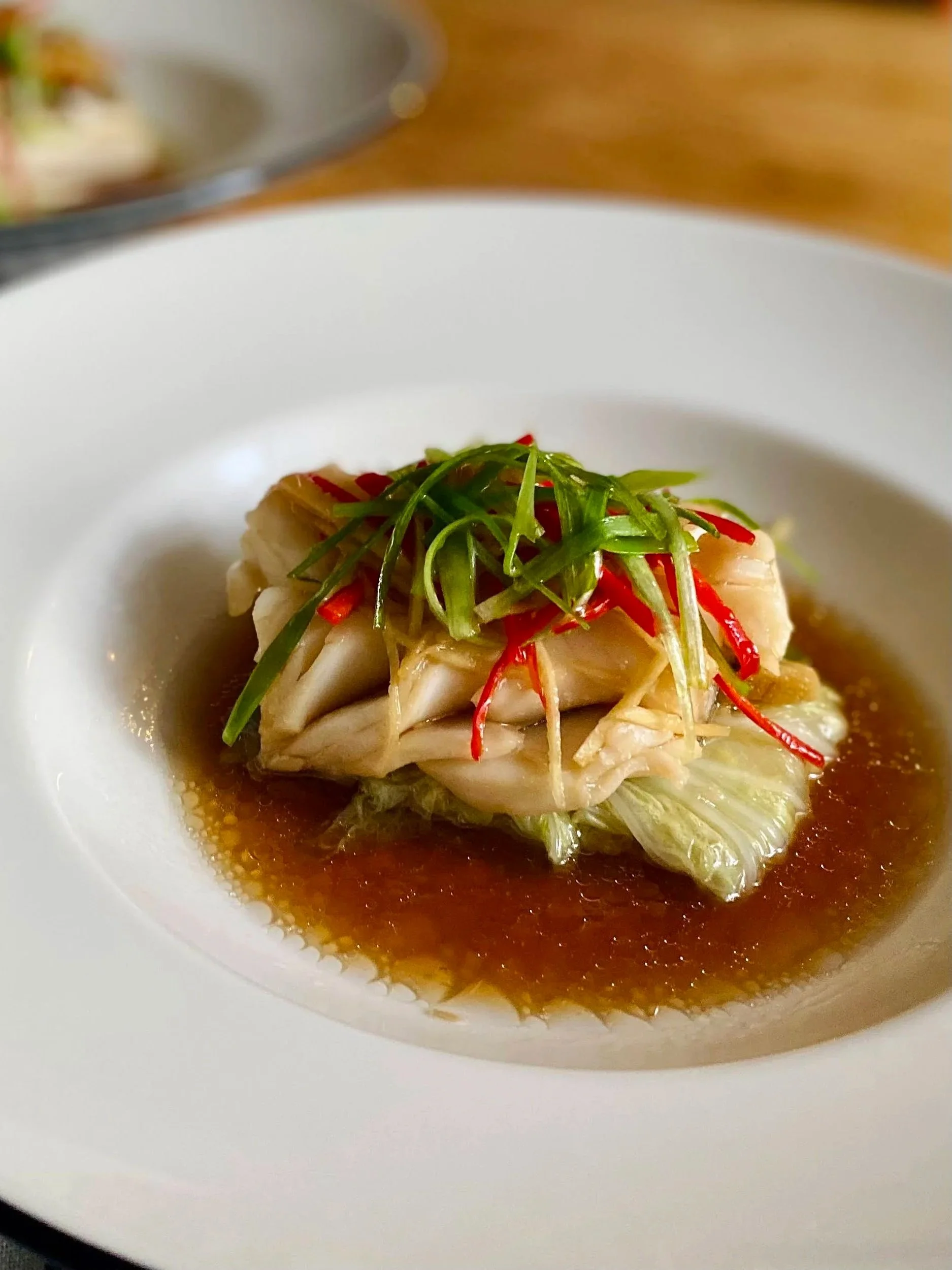 A plated dish with a piece of fish topped with shredded green and red peppers, served in a savory broth.
