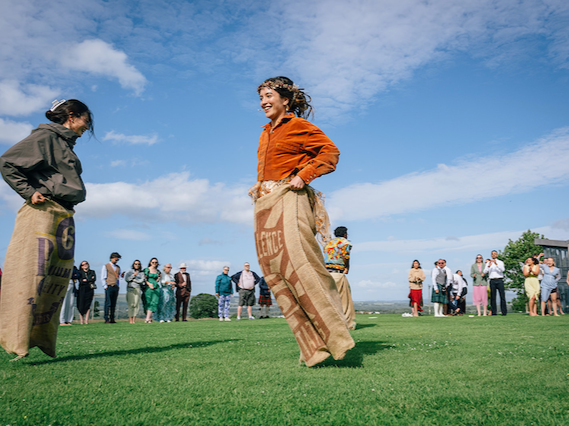 People participating in a sack race outdoors on a grassy field with a blue sky and clouds.