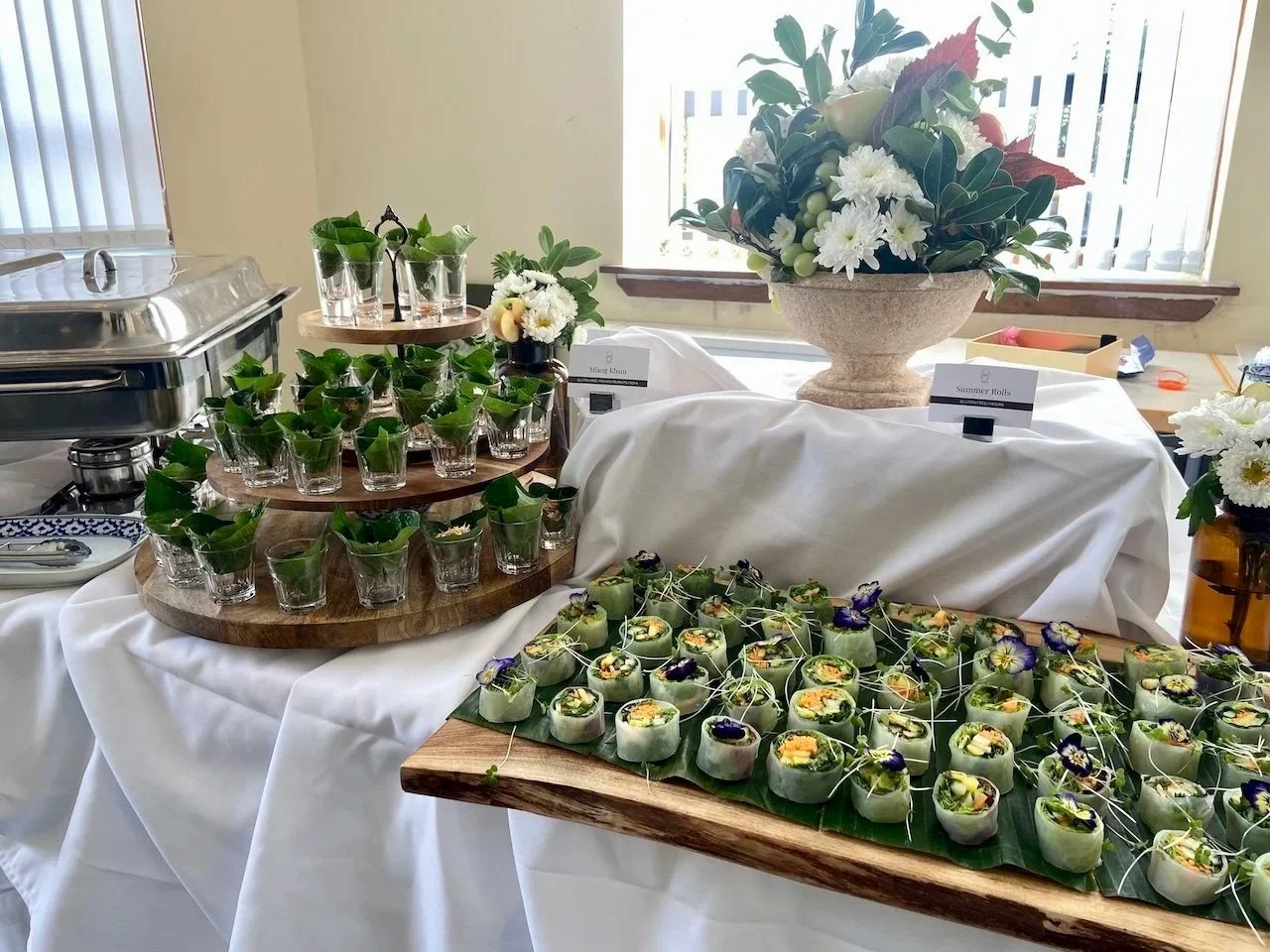 A buffet table with green wrapped appetizers, a large floral centerpiece, and small glasses with green leaves, set in a bright room with windows.