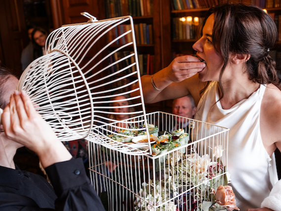 A woman in a white dress is feeding herself sushi from a large white wire basket at a gathering in a cozy, wood-lined room with bookshelves, while others watch and smile.