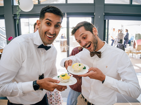 Two smiling men in formal attire holding plates with small desserts, celebrating indoors.