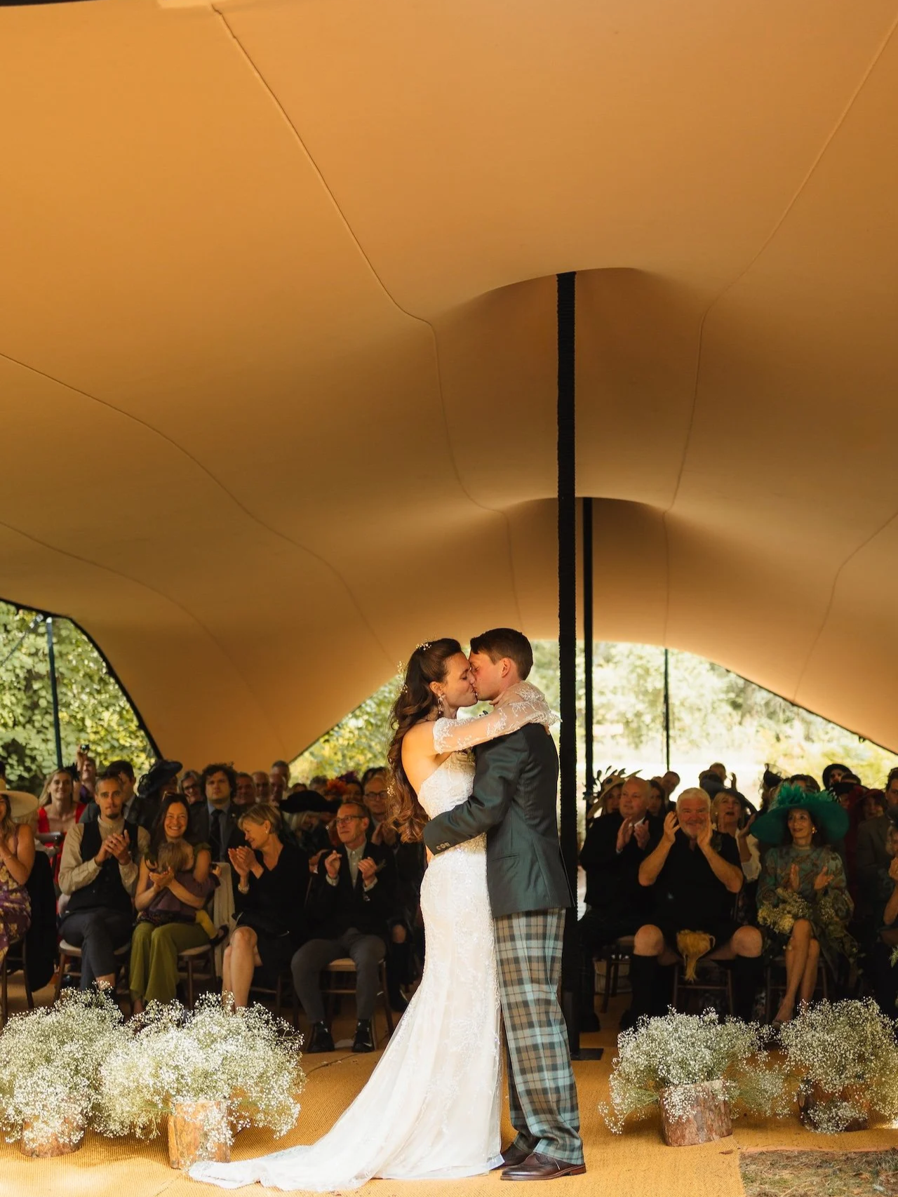 A bride and groom share a kiss during their wedding ceremony underneath a modern, curved ceiling with an audience watching.