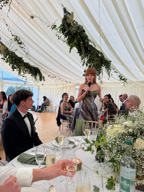 A woman is giving a speech at a wedding reception inside a decorated tent. The bride and groom sit at a table with drinks and floral arrangements. The tent is adorned with hanging greenery and string lights.