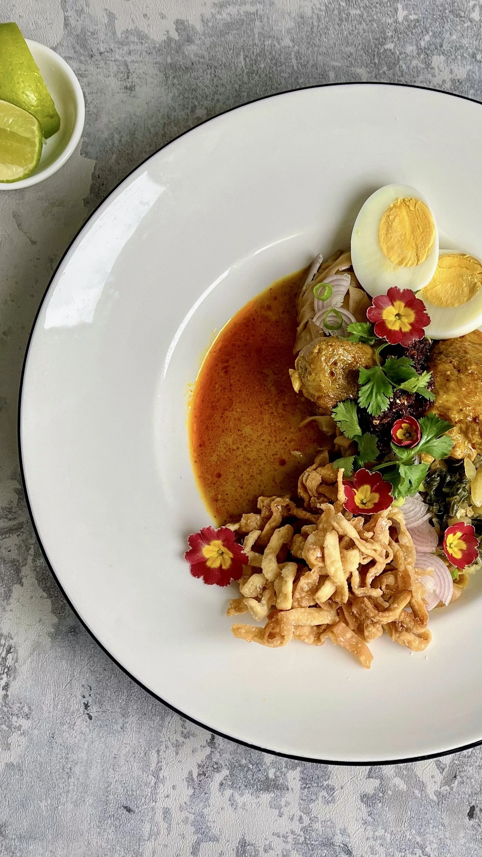 Plate of Thai curry with boiled eggs, fried noodles, garnished with cilantro, edible flowers, and sliced onions, accompanied by lime wedges.