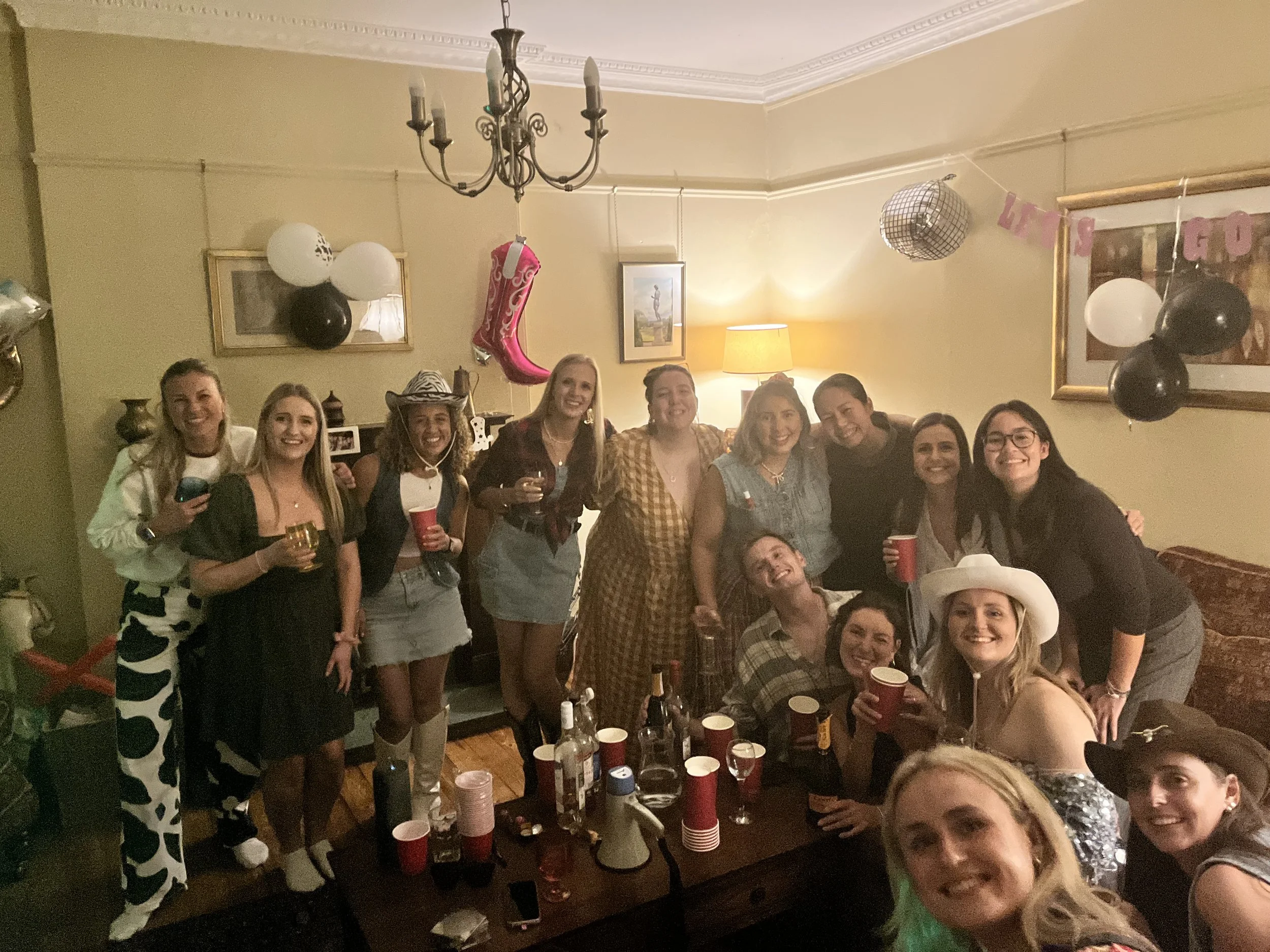A group of women at a party, smiling and holding drinks, in a decorated living room with balloons, a disco ball, and a banner.