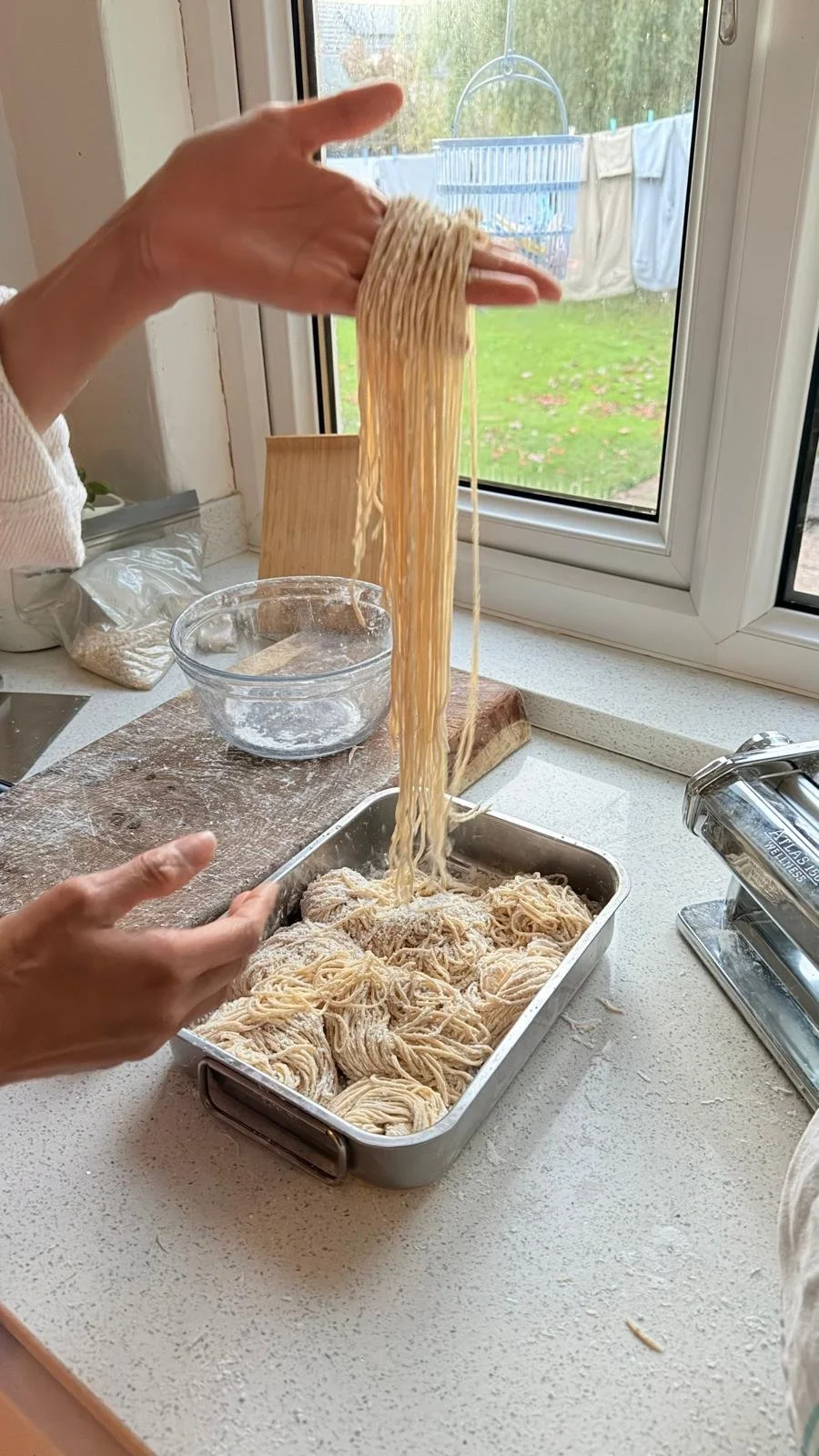 Person lifting freshly made spaghetti noodles from a baking dish in a kitchen near a window.
