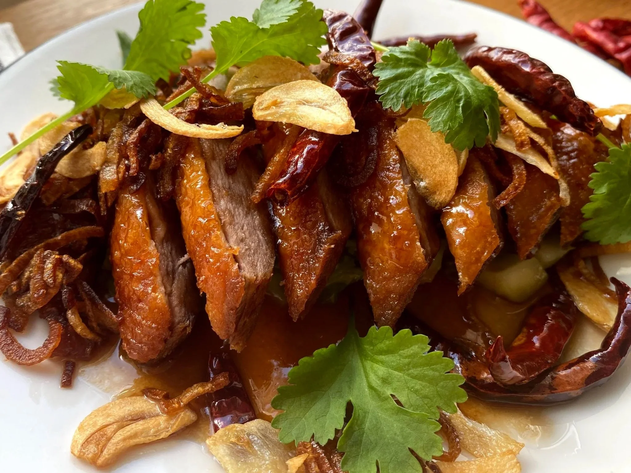 Close-up of an Asian-style roasted meat dish garnished with cilantro, fried garlic, dried red chilies, and crispy shallots