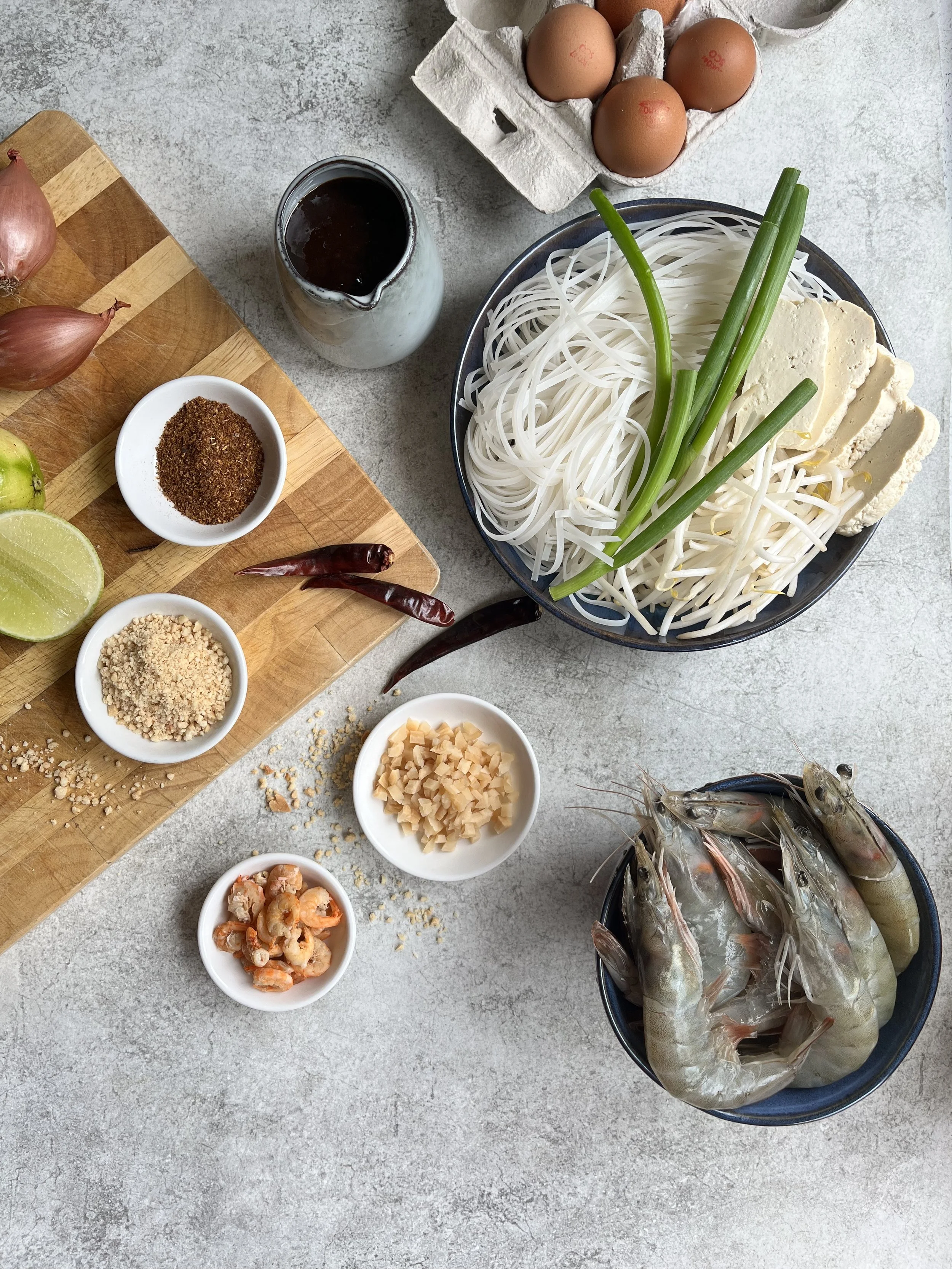 Ingredients for a dish, including shrimp, rice noodles, tofu, eggs, lime, dried chili peppers, crushed peanuts, chopped garlic, and various seasonings, arranged on a kitchen surface.