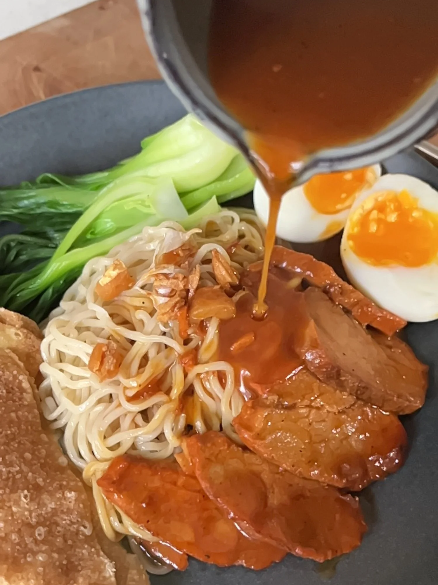 Dish with noodles, bok choy, two soft-boiled eggs, barbecue pork slices, fried chicken, and broth being poured over the noodles.