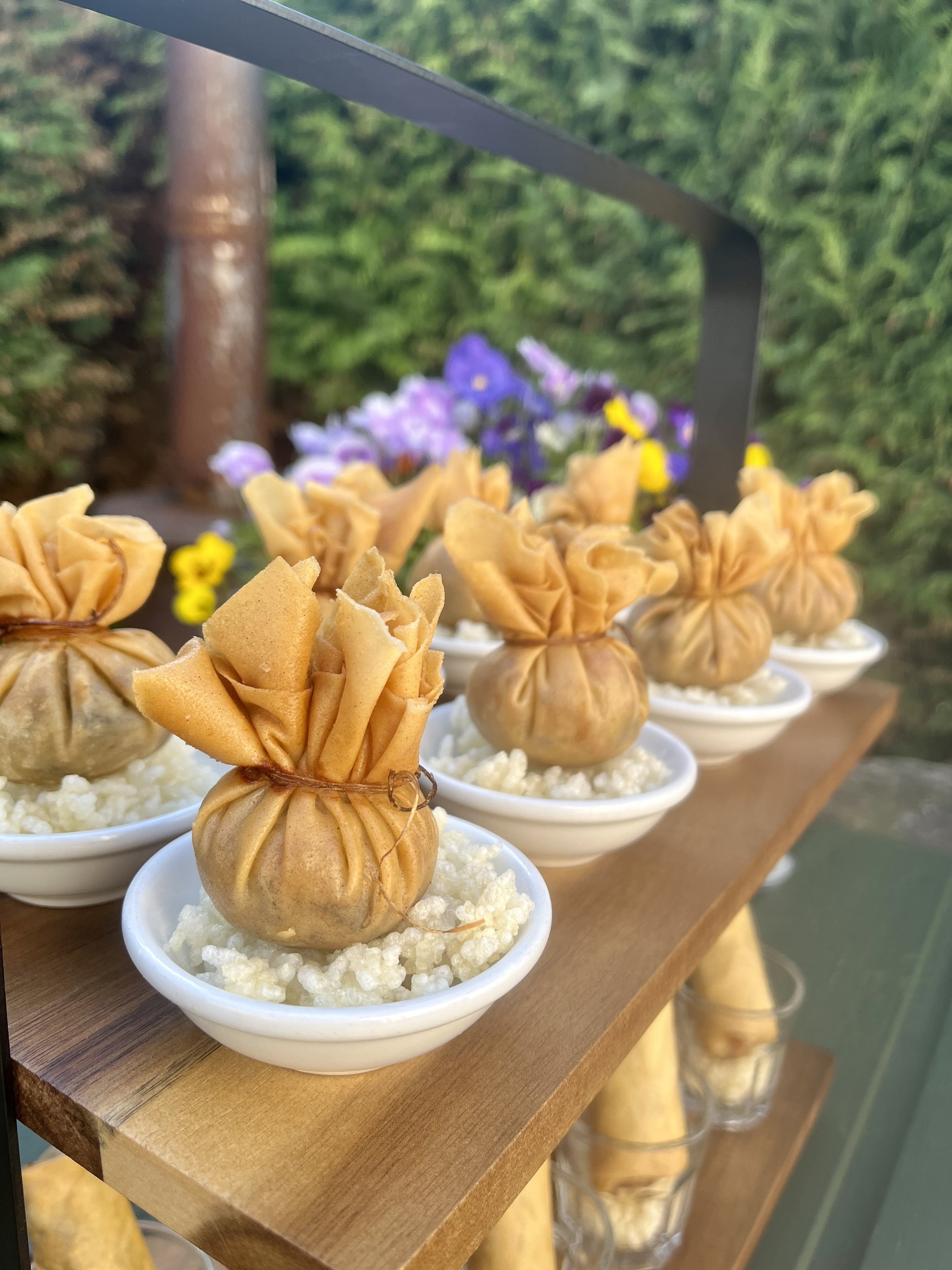 Small white bowls filled with white rice or grains, topped with fried plantains shaped like small pouches, arranged on a wooden tray with purple and yellow flowers in the background.