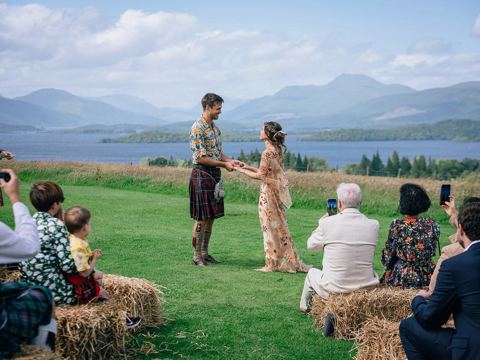 A wedding ceremony outdoors with a scenic lake and mountains in the background. The couple is holding hands and exchanging vows, with guests sitting on hay bales watching and taking photos.