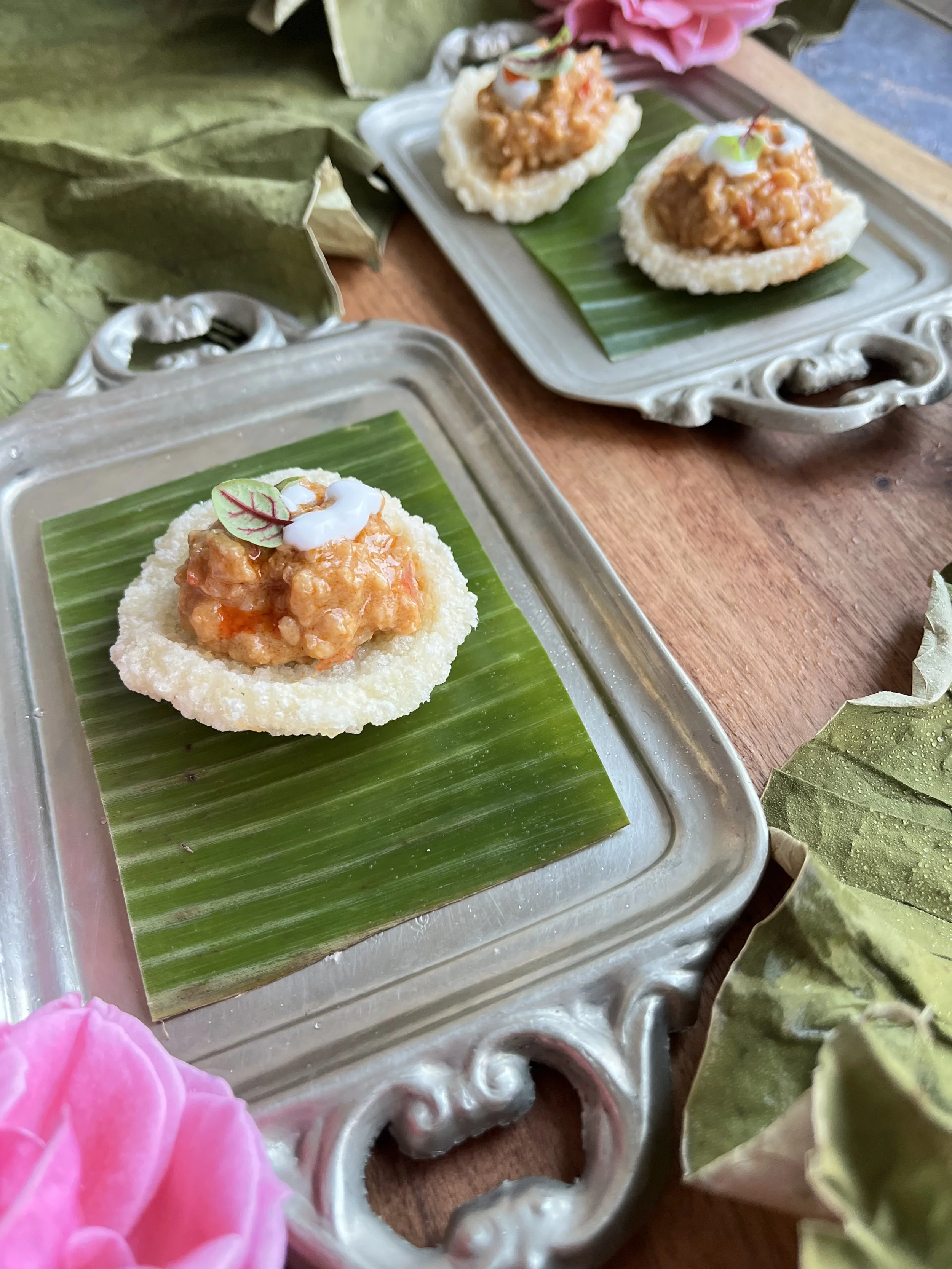 Two appetizer bite-sized servings on decorative silver trays with ornate edges, each on a green banana leaf. The servings contain a crispy fried cup filled with a spicy, chunky meat mixture, topped with a dollop of white sauce and microgreens. The trays are on a wooden surface decorated with pink flowers and dried leaves.