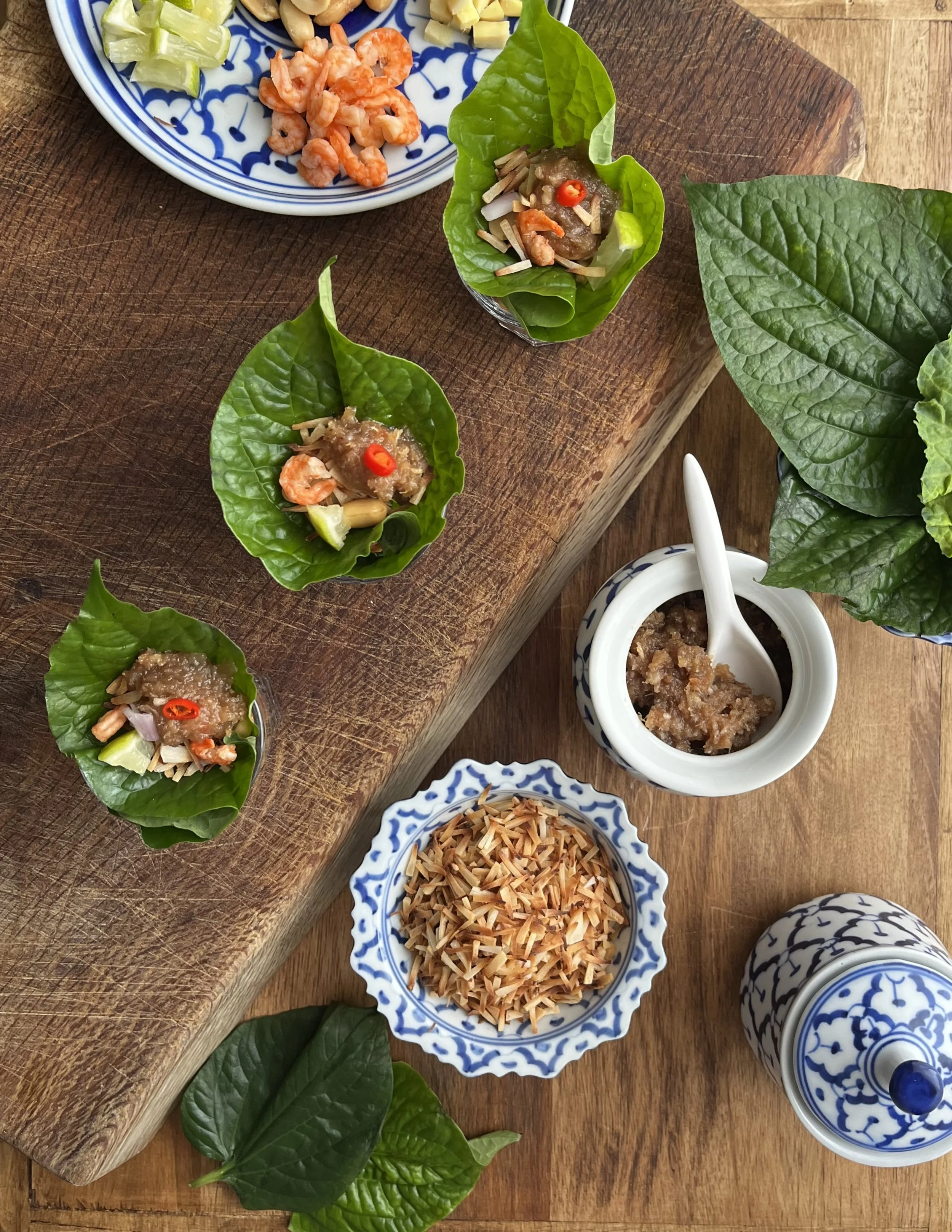 Three small leaf cups with shrimp and sauce, a bowl of shredded toasted coconut, a container of fermented shrimp paste, and a bowl of sliced dried ginger, all arranged on a wooden surface.