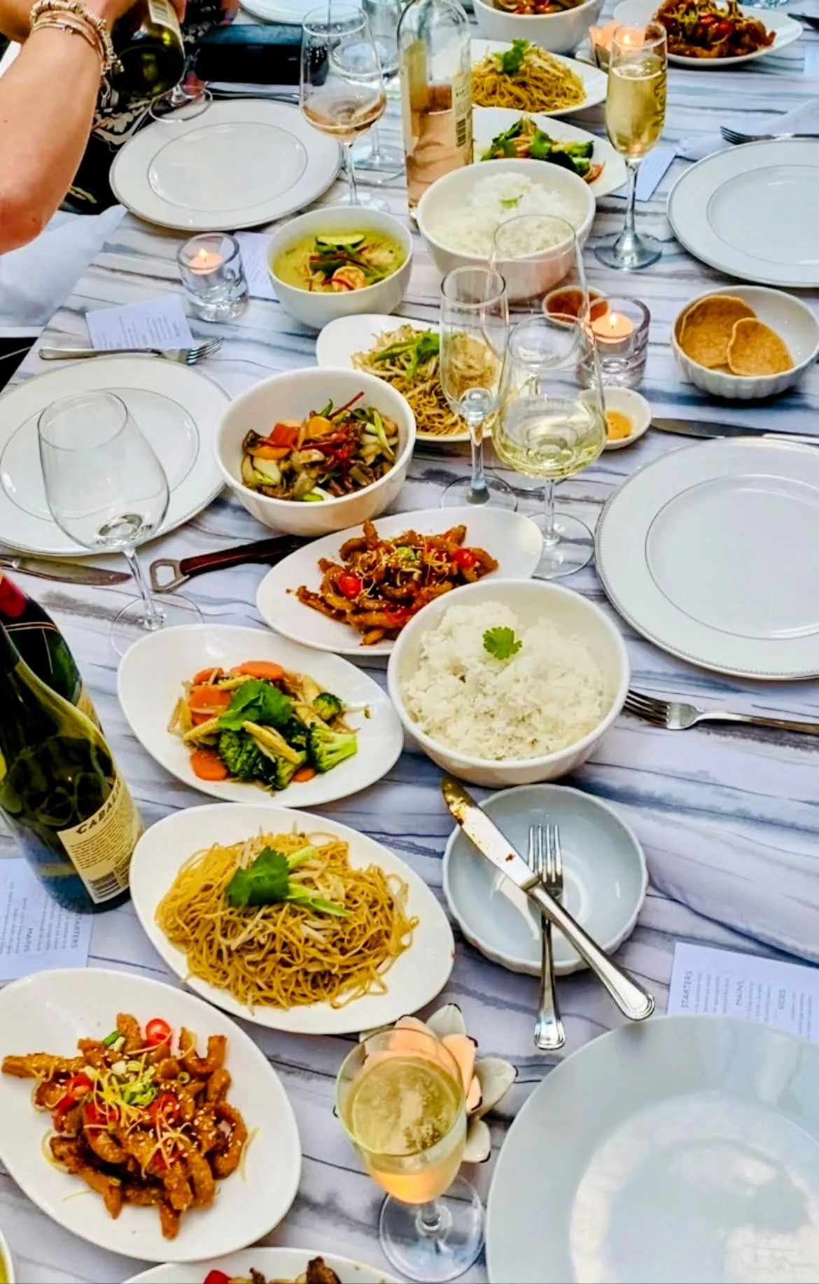 A table set for a meal with various Asian dishes and beverages on it, including rice, noodles, vegetables, and fried foods, with wine, champagne, and a beer bottle.