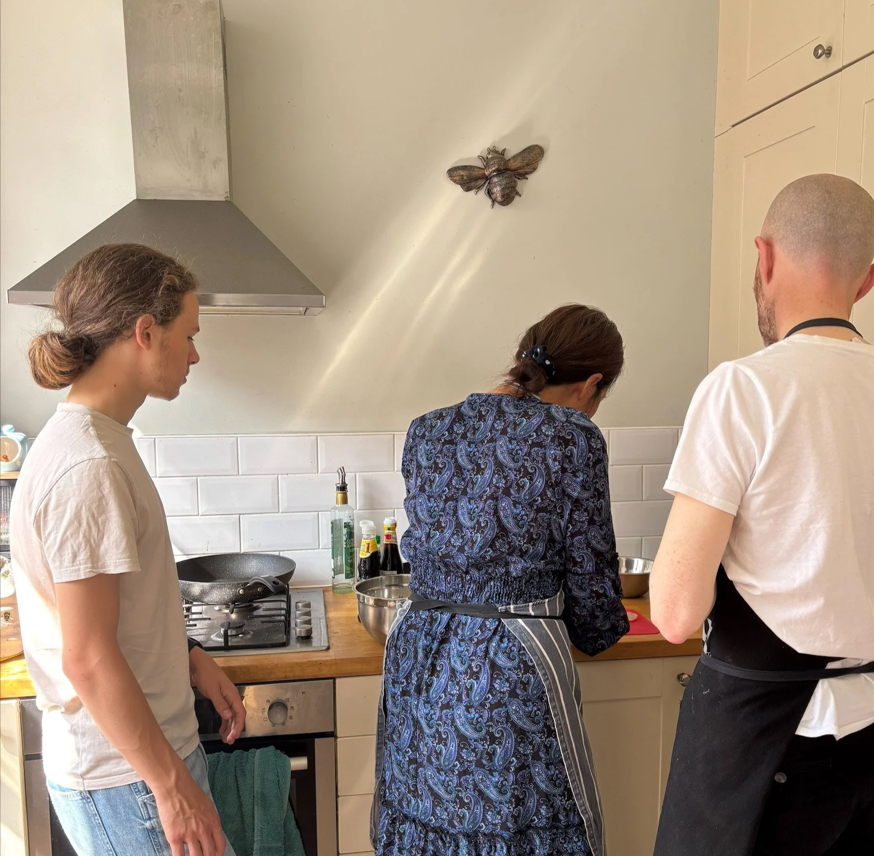 Three people cooking in a kitchen, two women and one man, with a bee decoration on the wall above them.