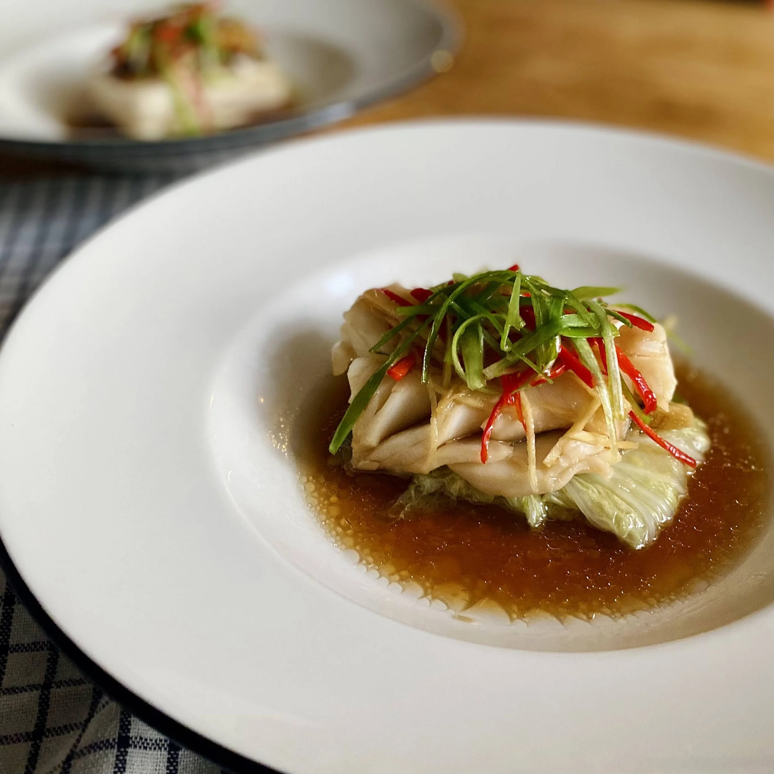 A plated Asian-style dumpling topped with shredded vegetables and green herbs, served with a soy-based sauce in a white dish.