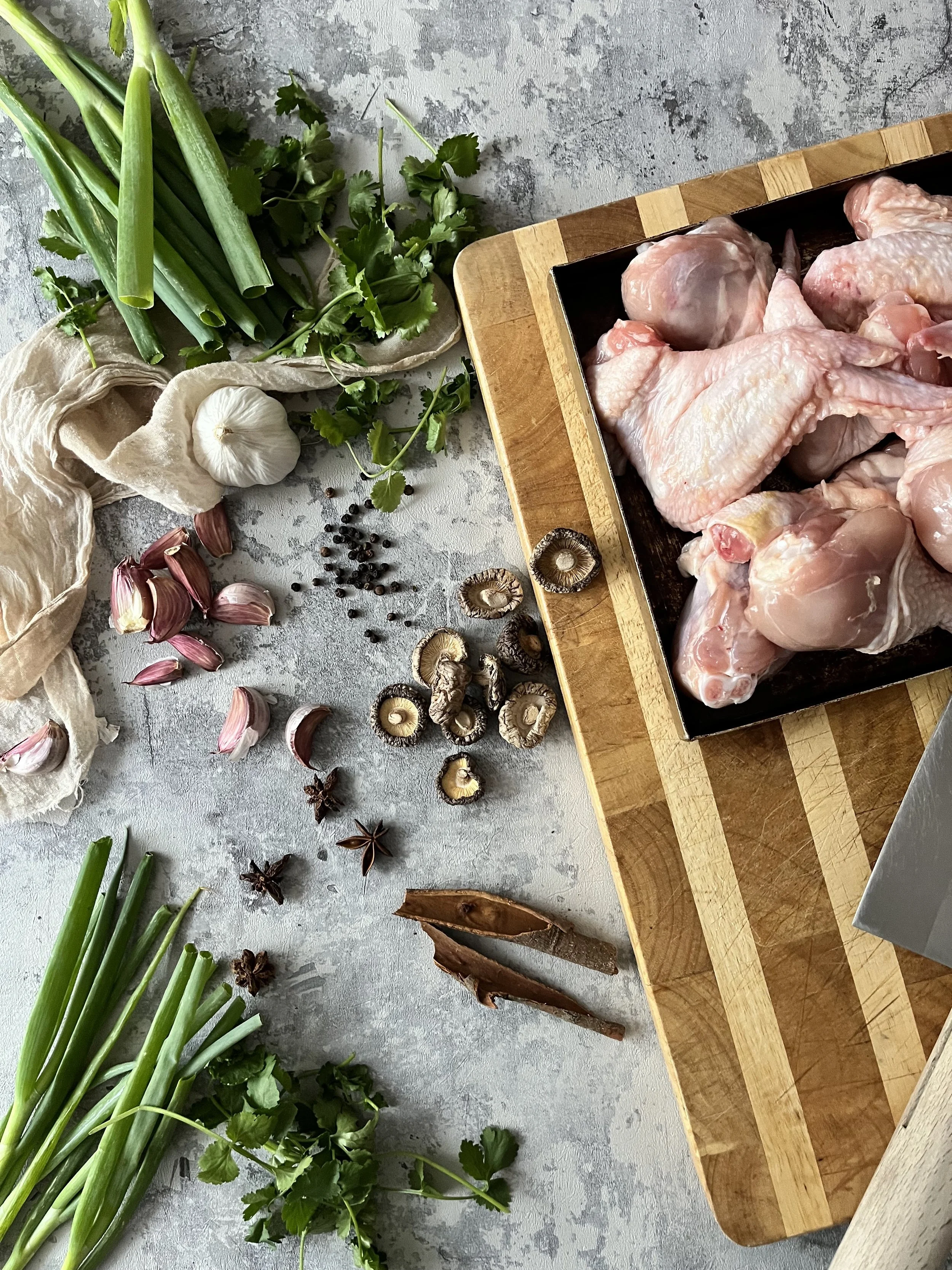 Raw chicken pieces on a cutting board surrounded by green onions, garlic, cilantro, dried mushrooms, star anise, garlic cloves, black peppercorns, shallots, and bay leaves.