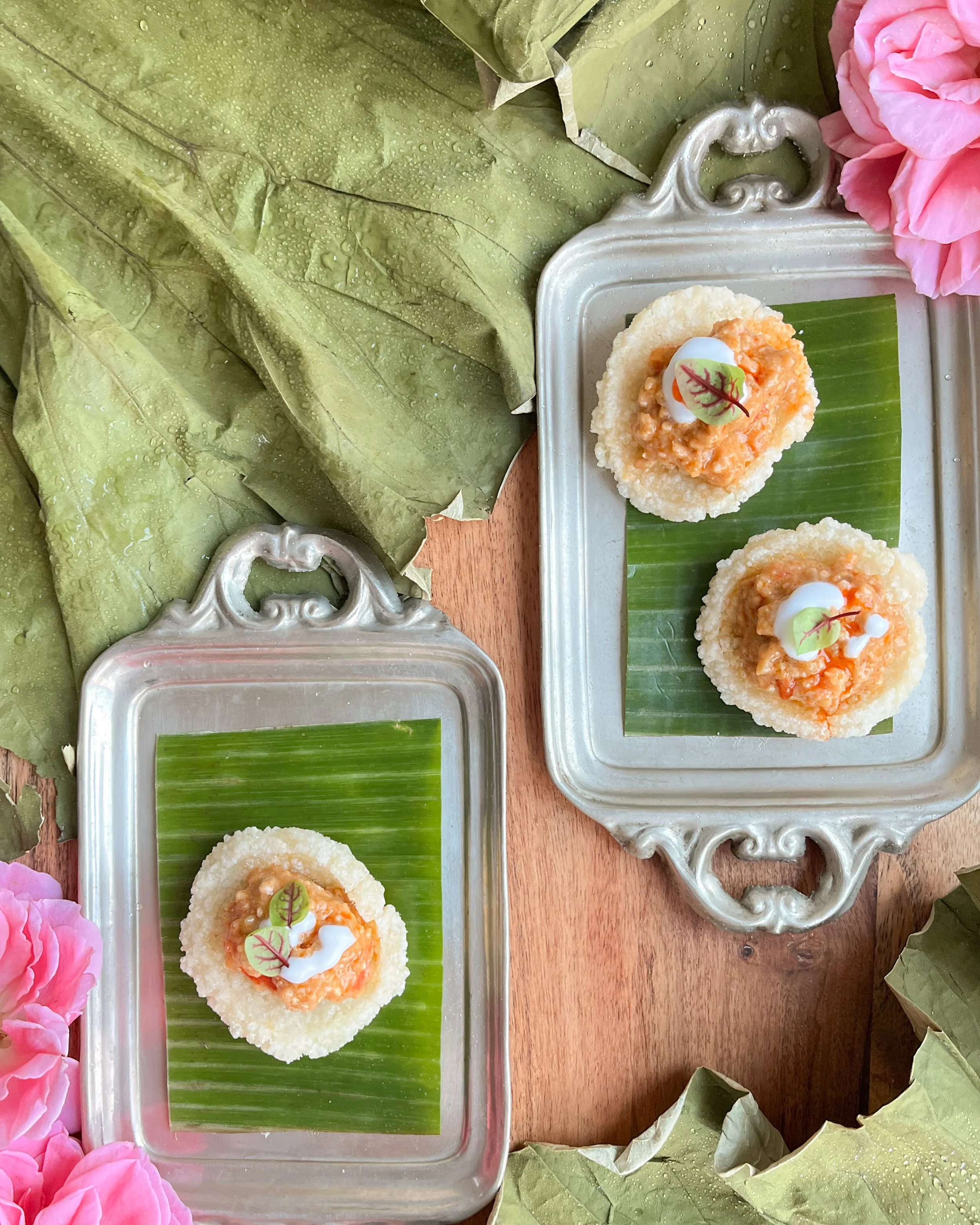 Two small appetizers on silver trays, each with a round crispy rice cracker topped with orange-colored spread, garnished with a dollop of white sauce and a small edible leaf, placed on a green banana leaf in a flower and leaf-decorated setting.