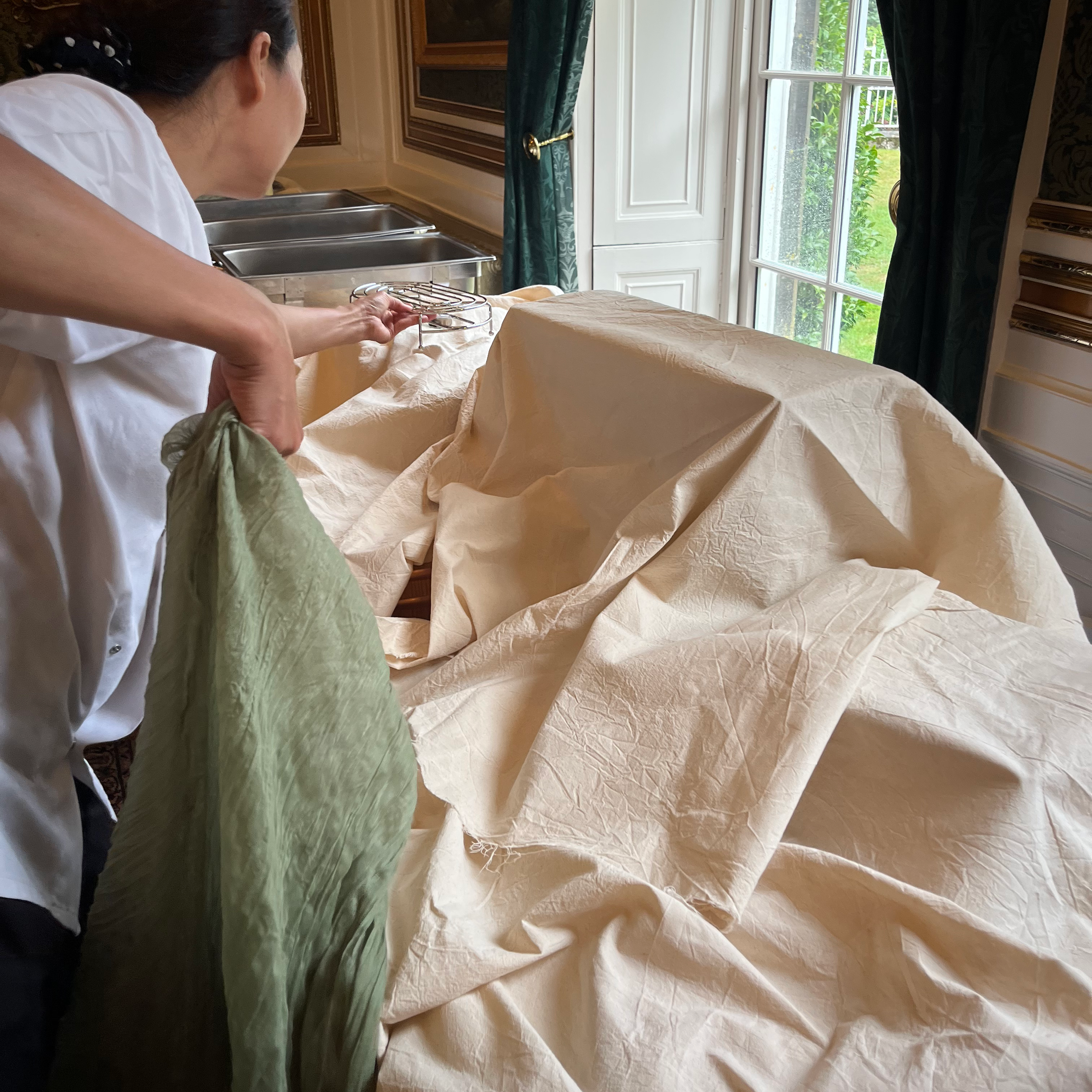 Person preparing to unveil an object covered with beige fabric in a well-lit room with large window, green curtain, and antique decor.