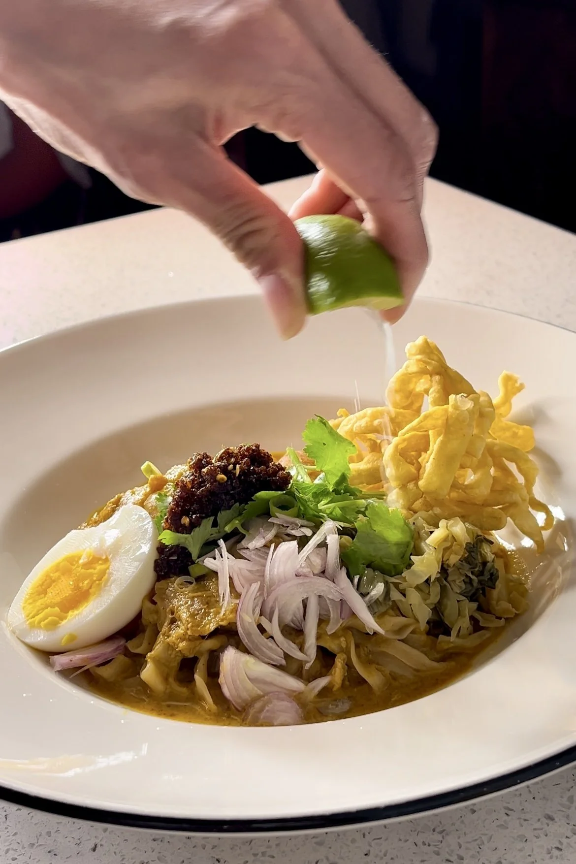 Person squeezing lime over a bowl of noodles topped with sliced boiled egg, onions, cilantro, and crispy fried strips.