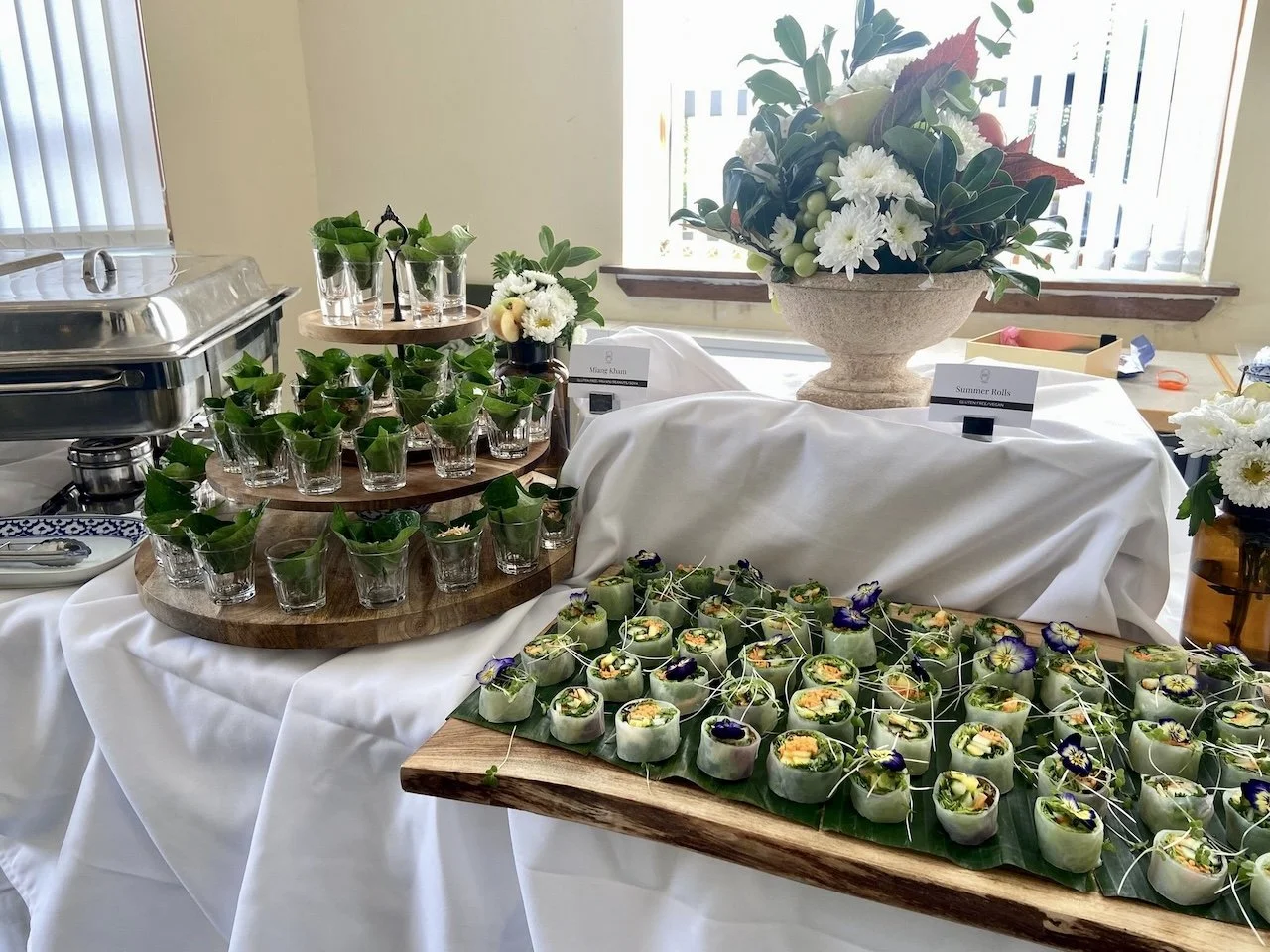 The image shows a beautifully arranged Thai wedding catering display, featuring appetizers seved in bai cha poo in the short glasss, a tiered stand with small bites and a flowers arrangement. 