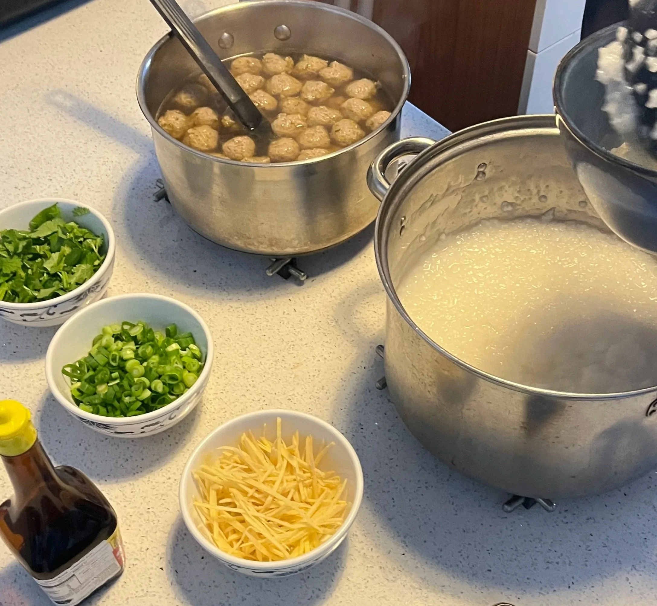 Pot of meatballs cooking in sauce, large pot of melted cheese, bowls of chopped green onions, cilantro, shredded cheese, a bottle of soy sauce, and a saucepan of rice on a kitchen counter.