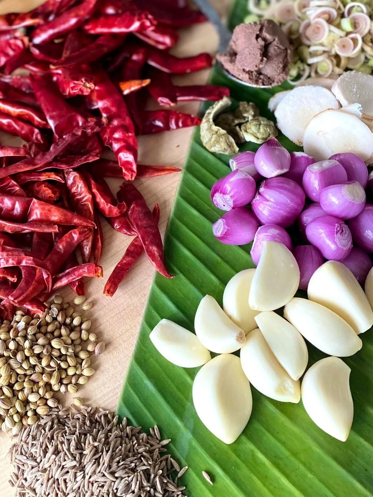 An assortment of colorful spices, garlic, shallots, dried chilies, and herbs arranged on a green banana leaf.