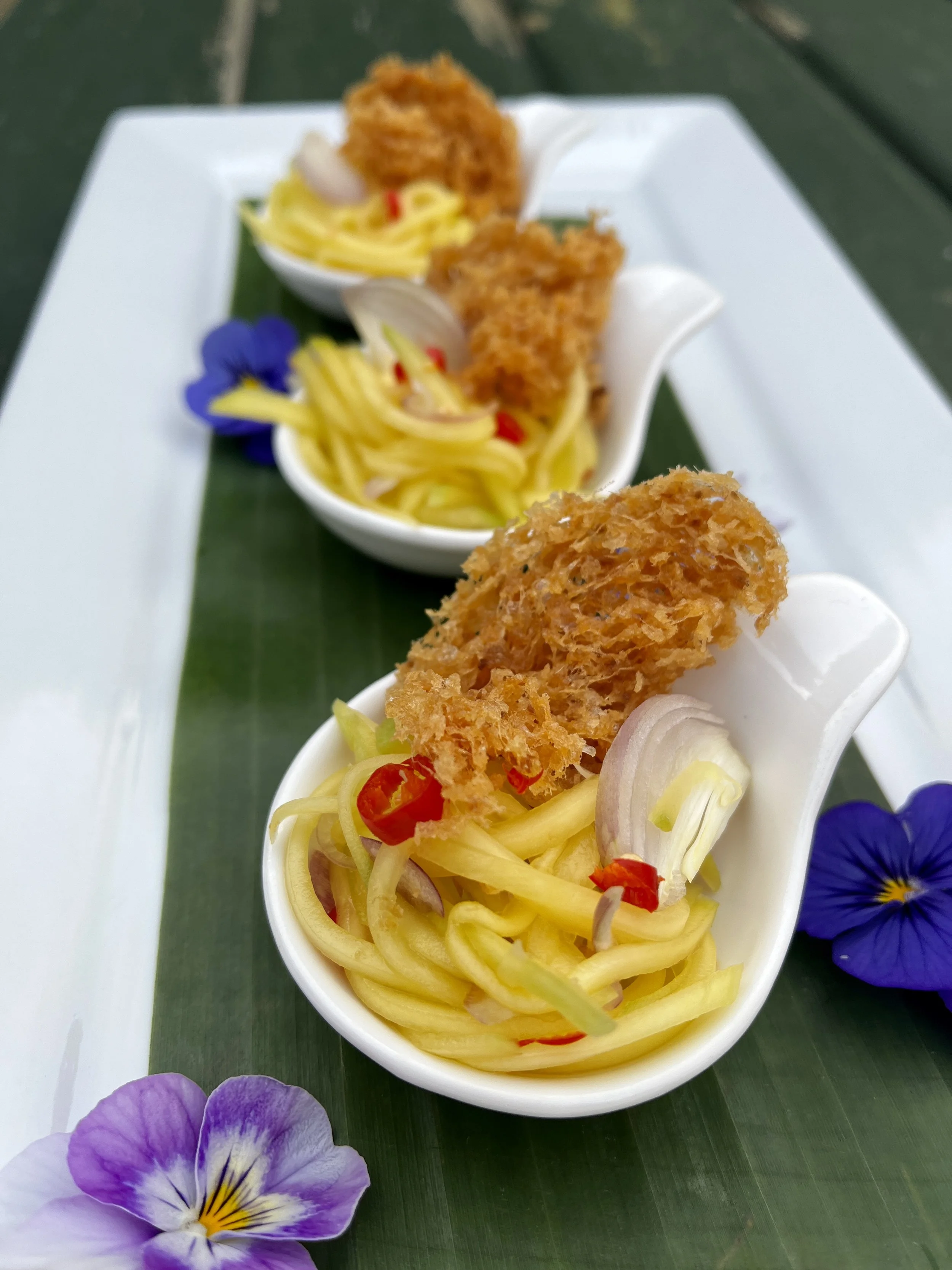 Three white ceramic dishes containing Asian-style noodle salad with fried fish skin, garnished with red chili slices, thinly sliced onions, and an edible purple flower, placed on a dark green leaf on a white rectangular plate.