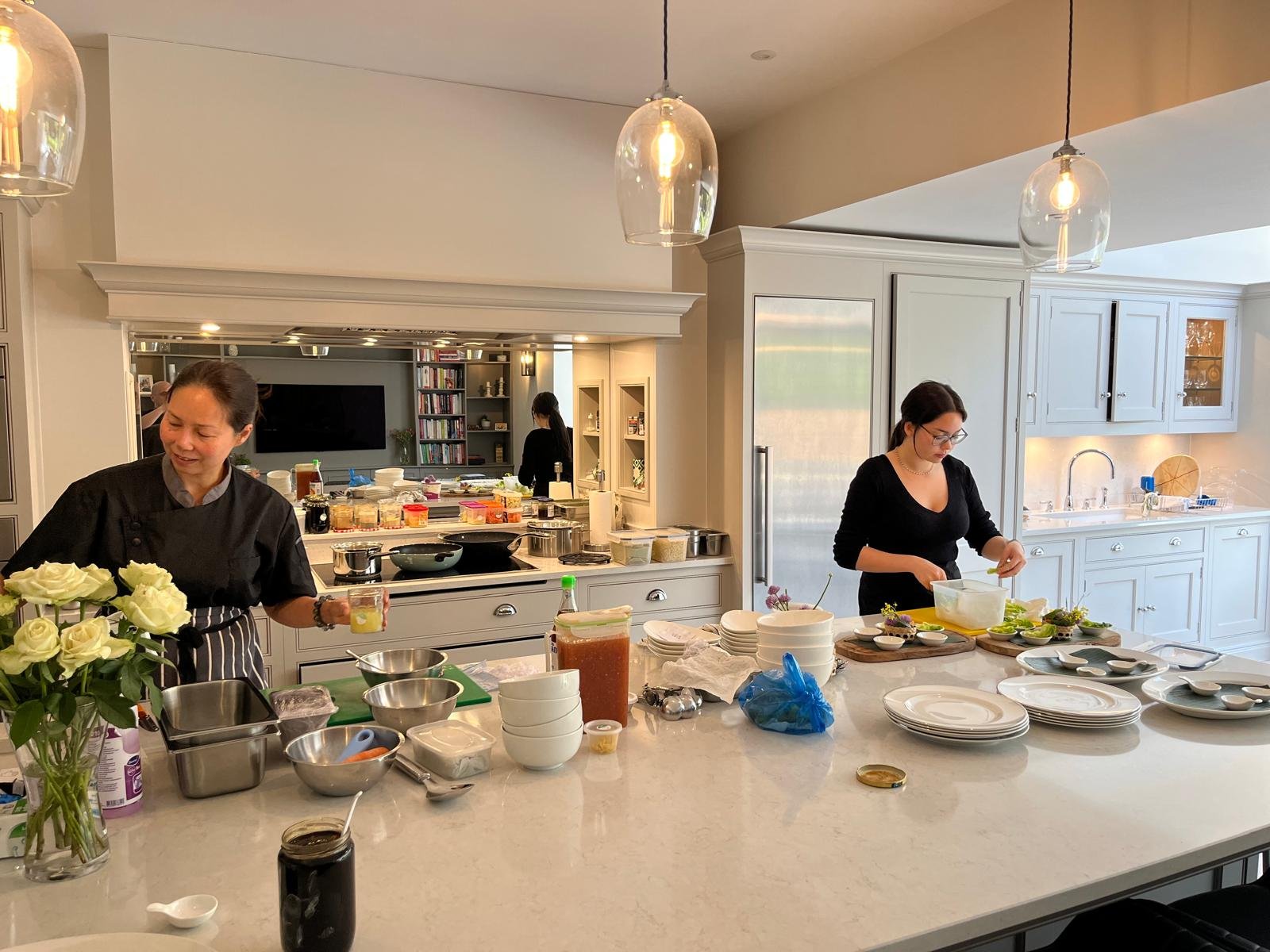 Two women preparing food in a modern kitchen with white cabinets and pendant lights, one woman with glasses and the other wearing a chef's coat.